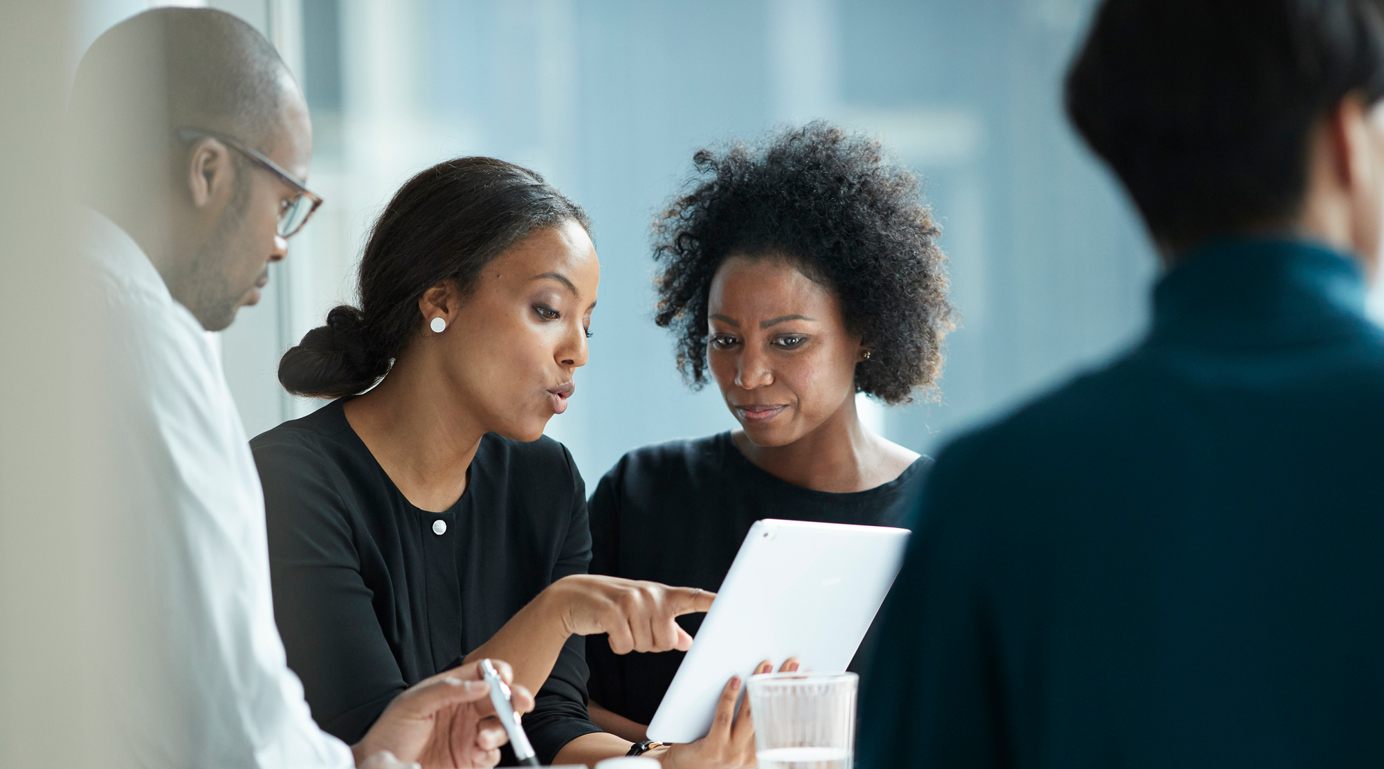 Four professionals in a business meeting observing a tablet, with focused expressions, sitting closely at a table in a modern office.