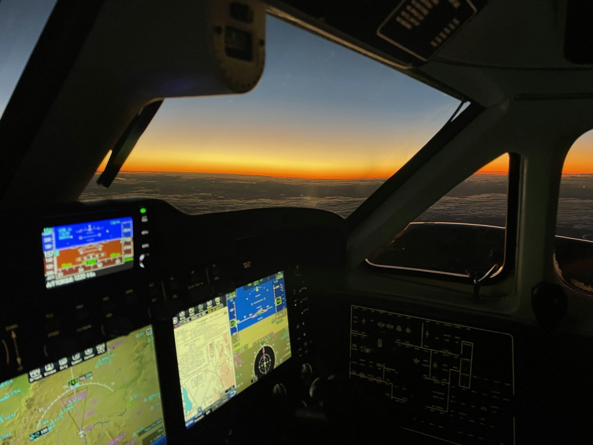 Inside the cockpit of an airplane showing multiple avionics screens displaying navigation information during a sunset or sunrise sky view through the front windows.