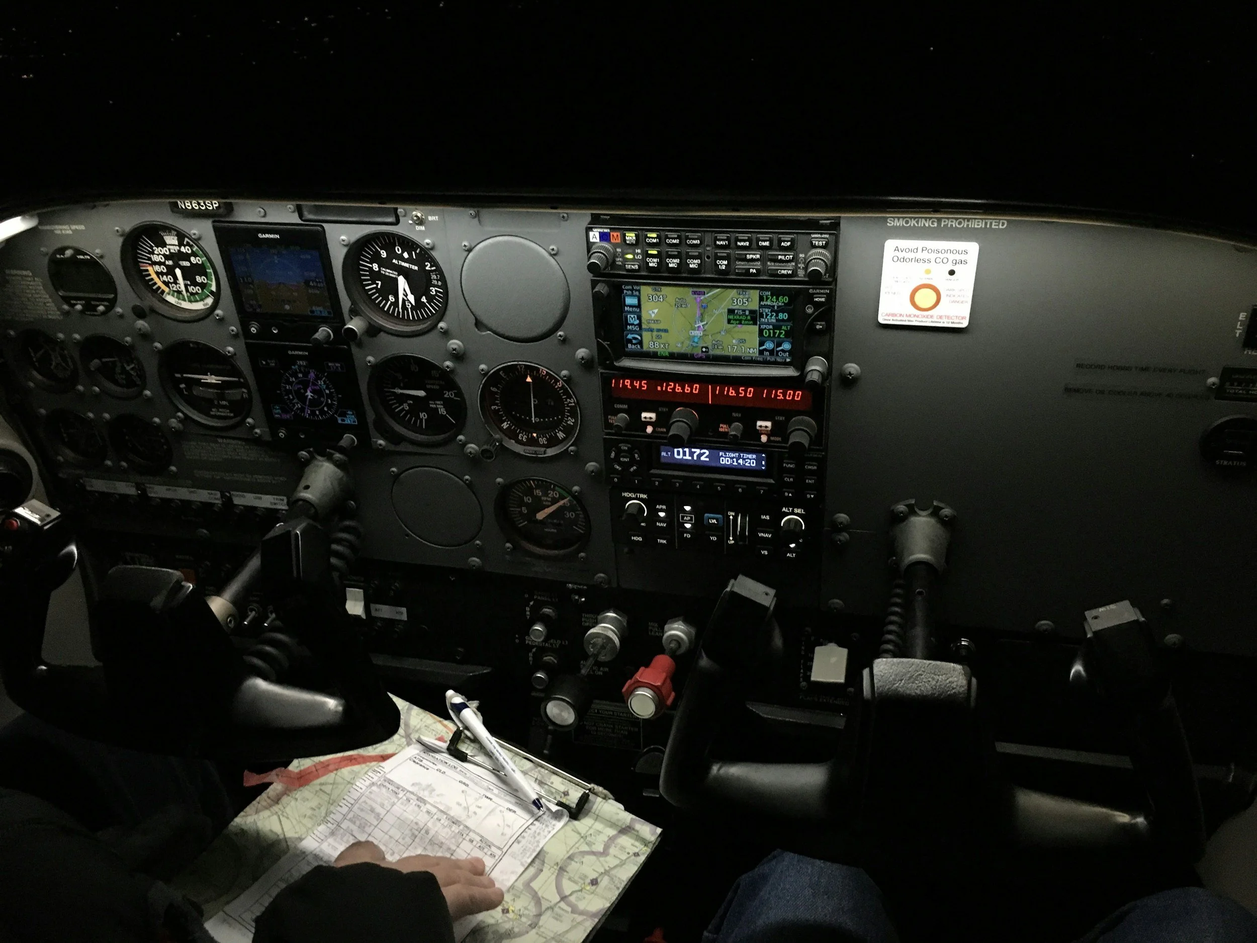 View of a small airplane cockpit with various instruments, gauges, and screens, and a pilot's hand writing on a flight log on a map.