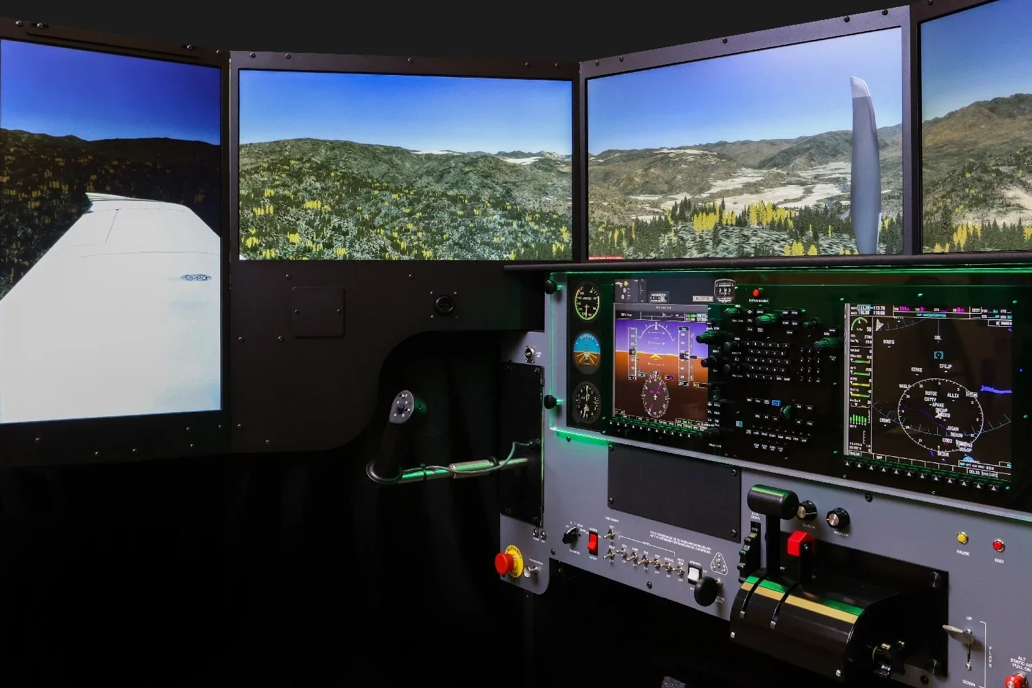 Inside an aircraft cockpit with multiple screens showing a mountainous landscape with trees and clear skies, control panels, and instruments.