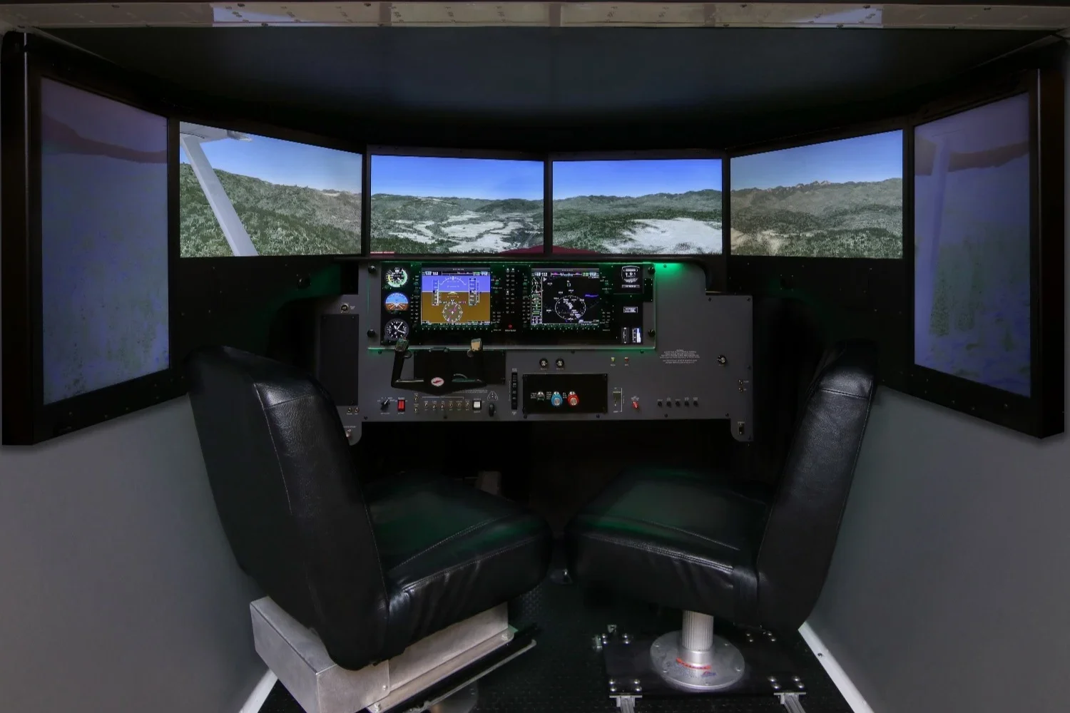 Inside an Redbird FMX flight simulator with three large screens displaying a landscape view of mountains and sky, with various control panels and two black leather seats.