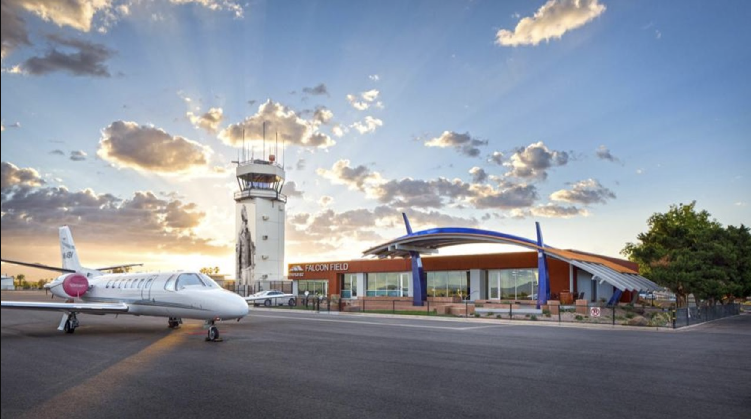 Small airport with an airplane on the tarmac, a control tower, and a modern building labeled Falcon Field during sunset. Trees and parked cars are visible in the background. KFFZ Falcon Field Airport, Mesa, Az.