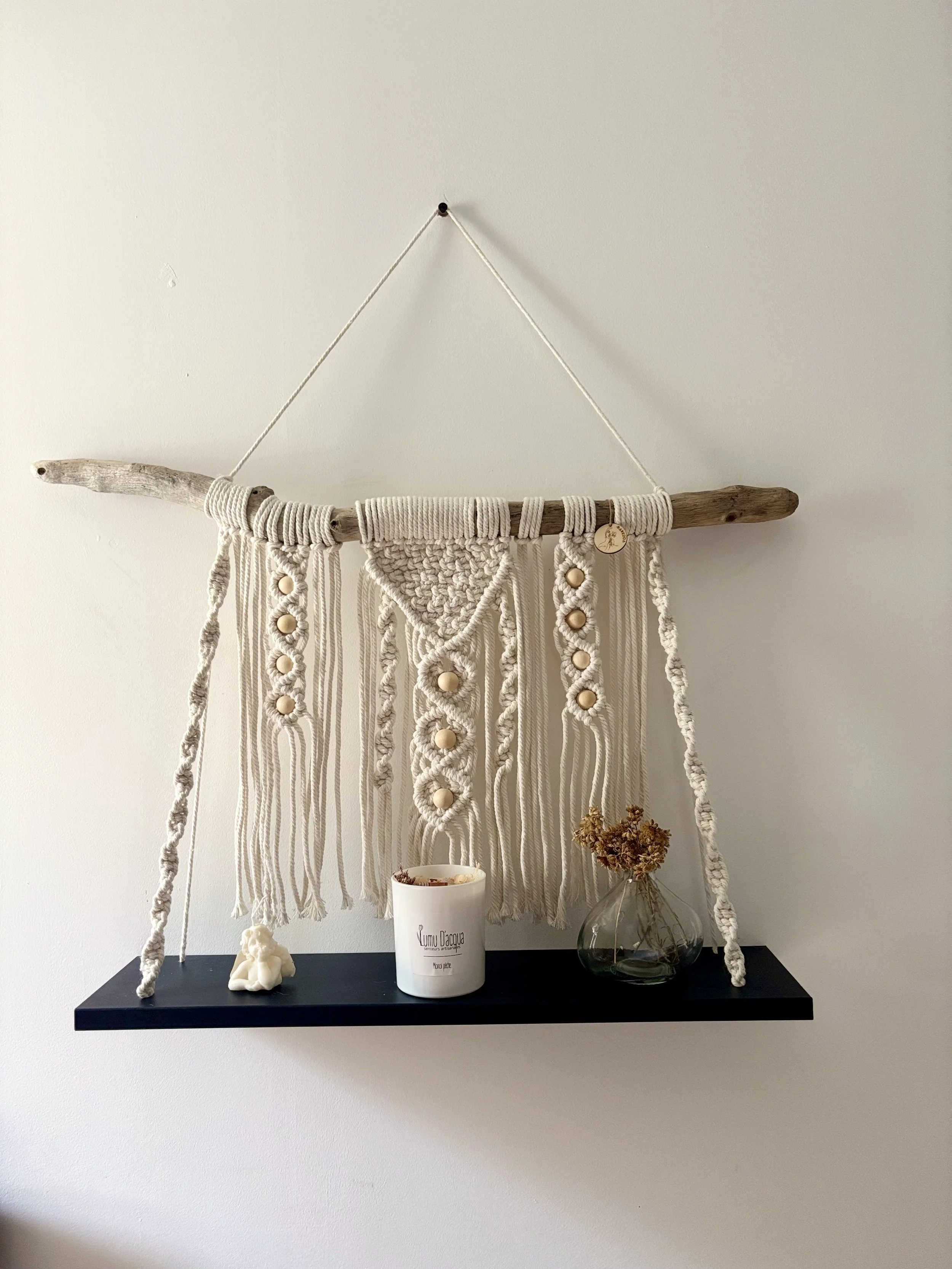 Une décoration murale en macramé suspendue à un morceau de bois, avec une petite figurine en cire, une bougie et un vase avec des fleurs séchées sur une étagère noire.