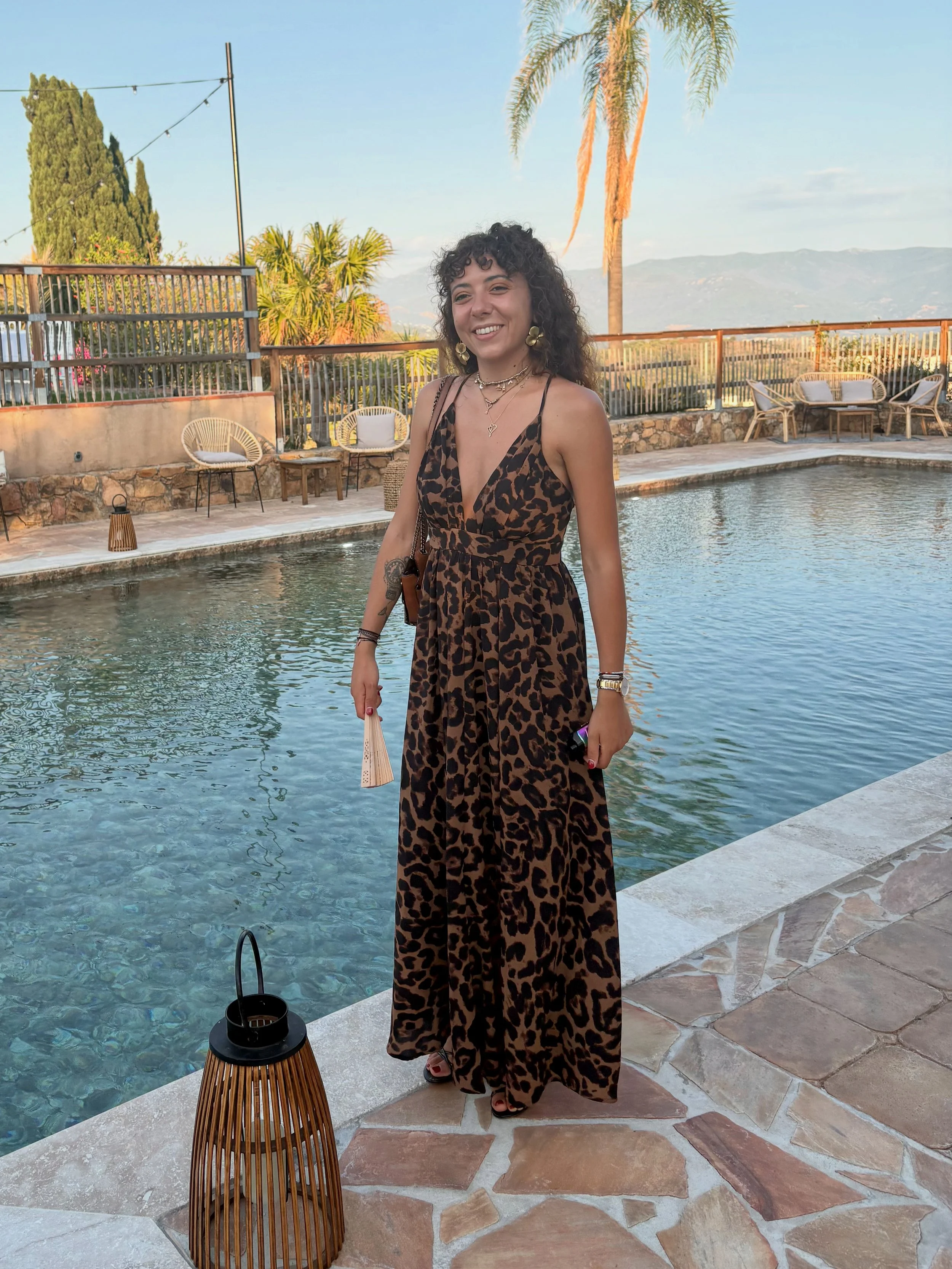 Jeune femme souriante en robe à motifs léopard debout près de la piscine avec décor tropical, palmiers, et mobilier d'extérieur