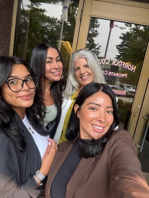 Five women taking a selfie outside of a building with glass doors, smiling and posing together.
