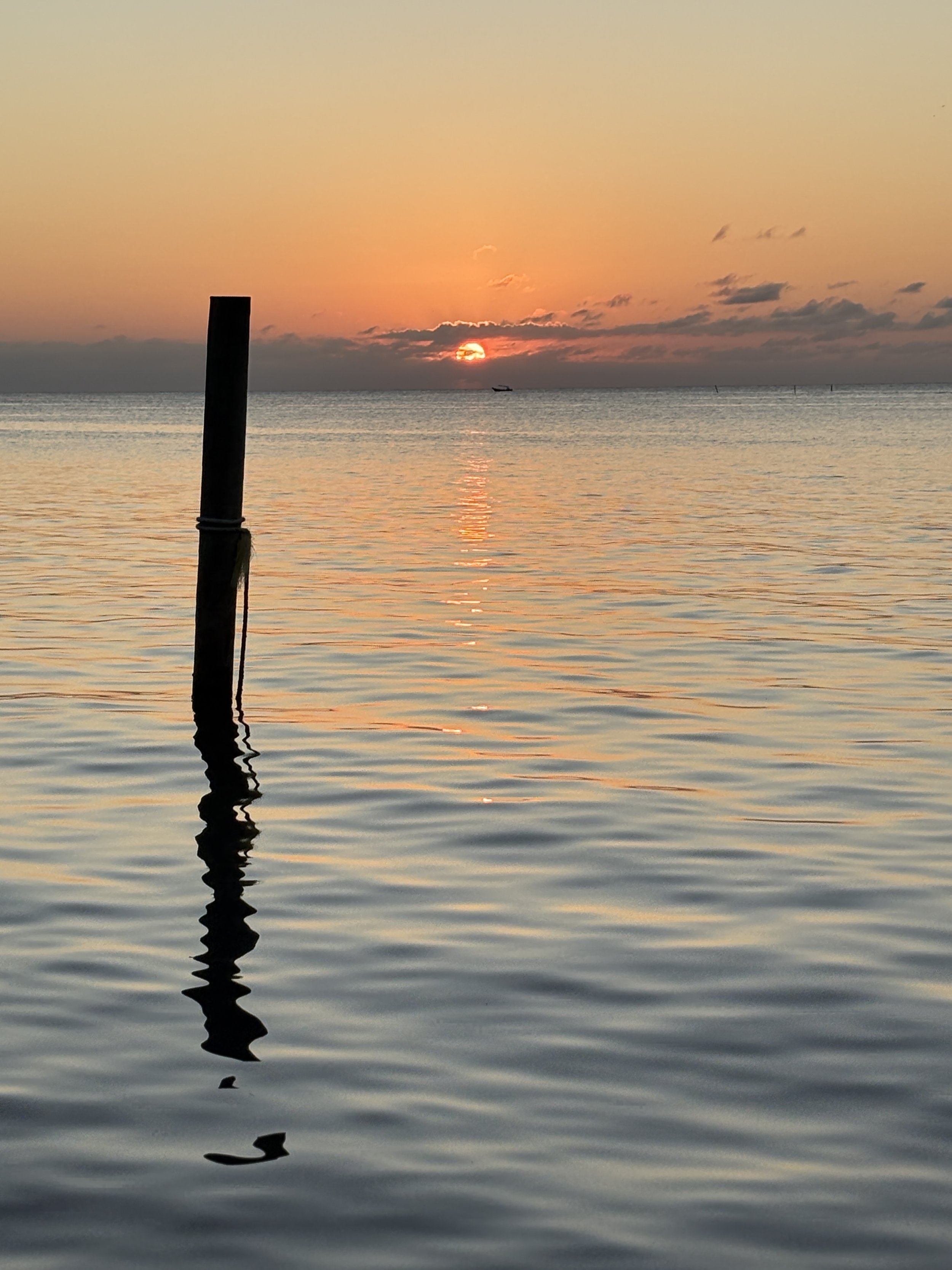 Sunset over a calm body of water with an orange and pink sky, a boat near the horizon, and a silhouette of a post reflected in the water.