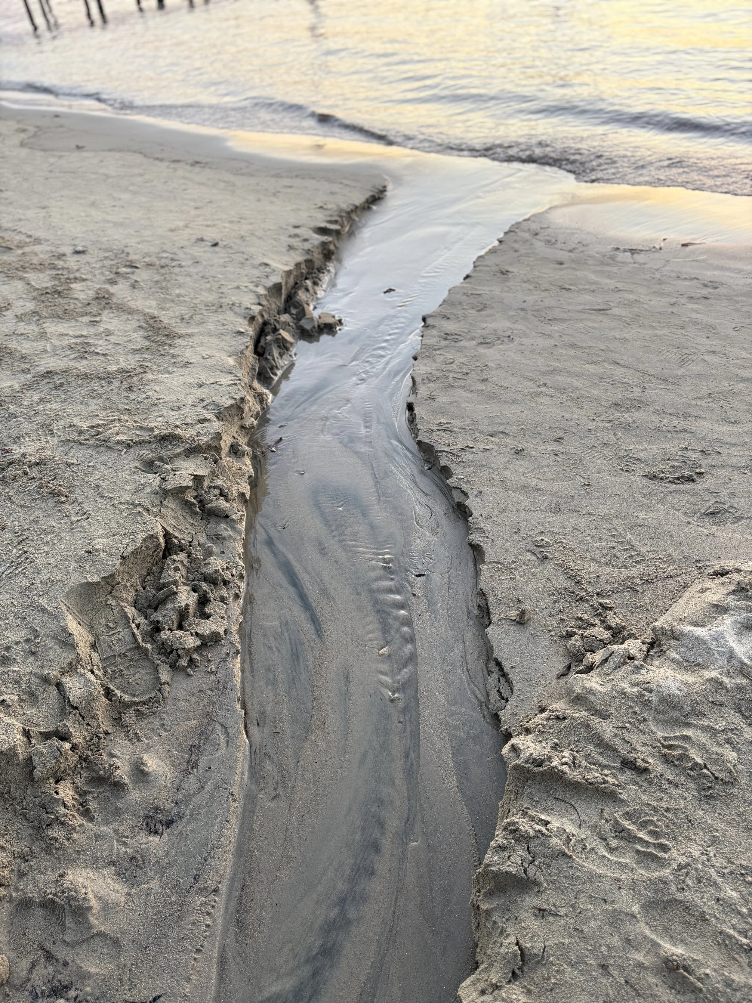 A small, narrow stream of water runs through the sand on a beach, with calm ocean waves in the background during sunset.