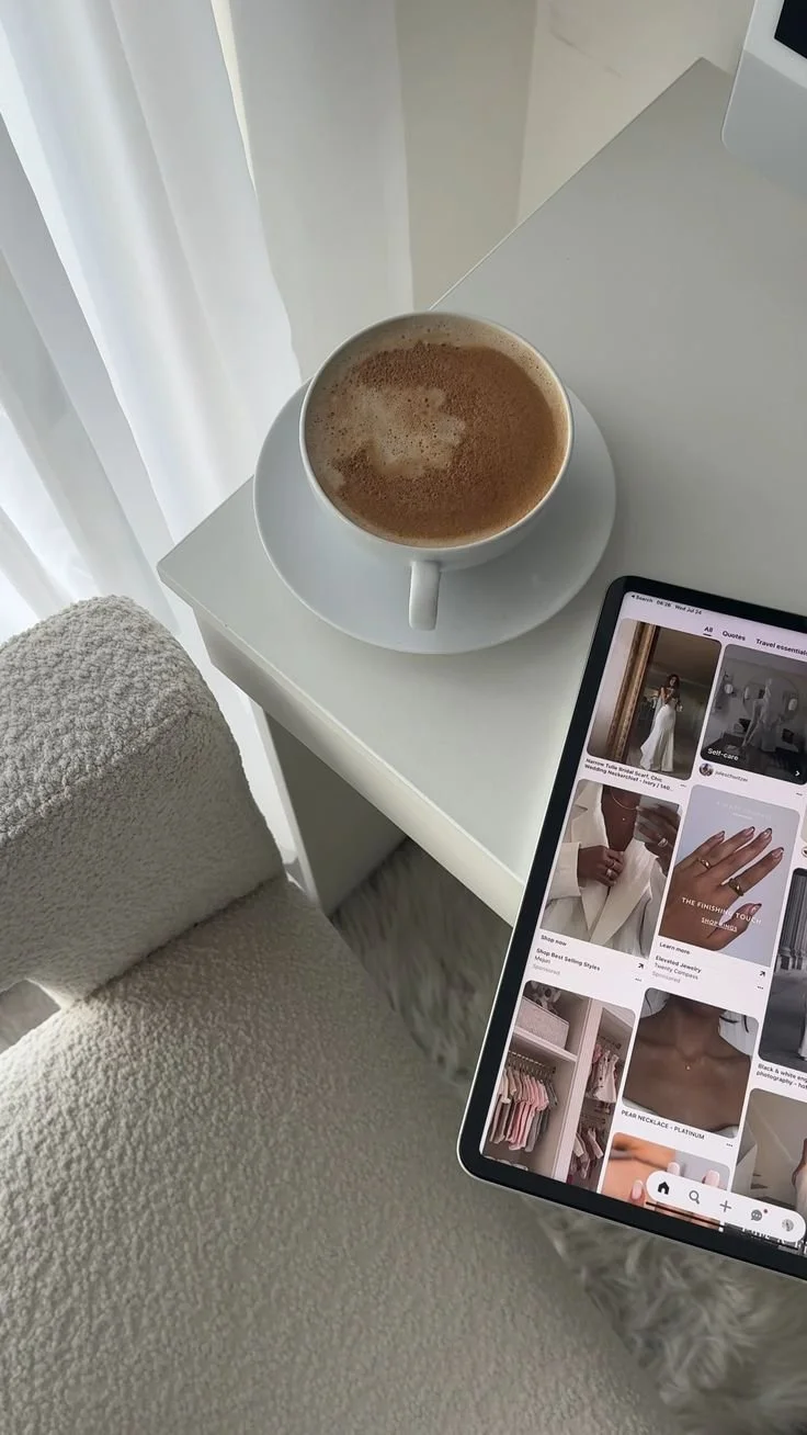 A cup of coffee on a saucer next to a tablet showing a fashion shopping website, on a white desk near a cozy beige chair and white curtains.