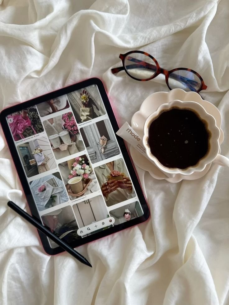 Tablet displaying a photo gallery, a pair of tortoiseshell glasses, a white flower-shaped cup of black coffee, a black stylus, and a white box of tea on a white cloth background.