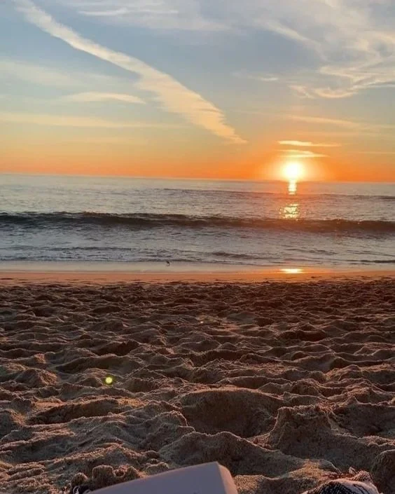 Sunset over the ocean seen from a sandy beach with footprints, with wispy clouds in the sky.