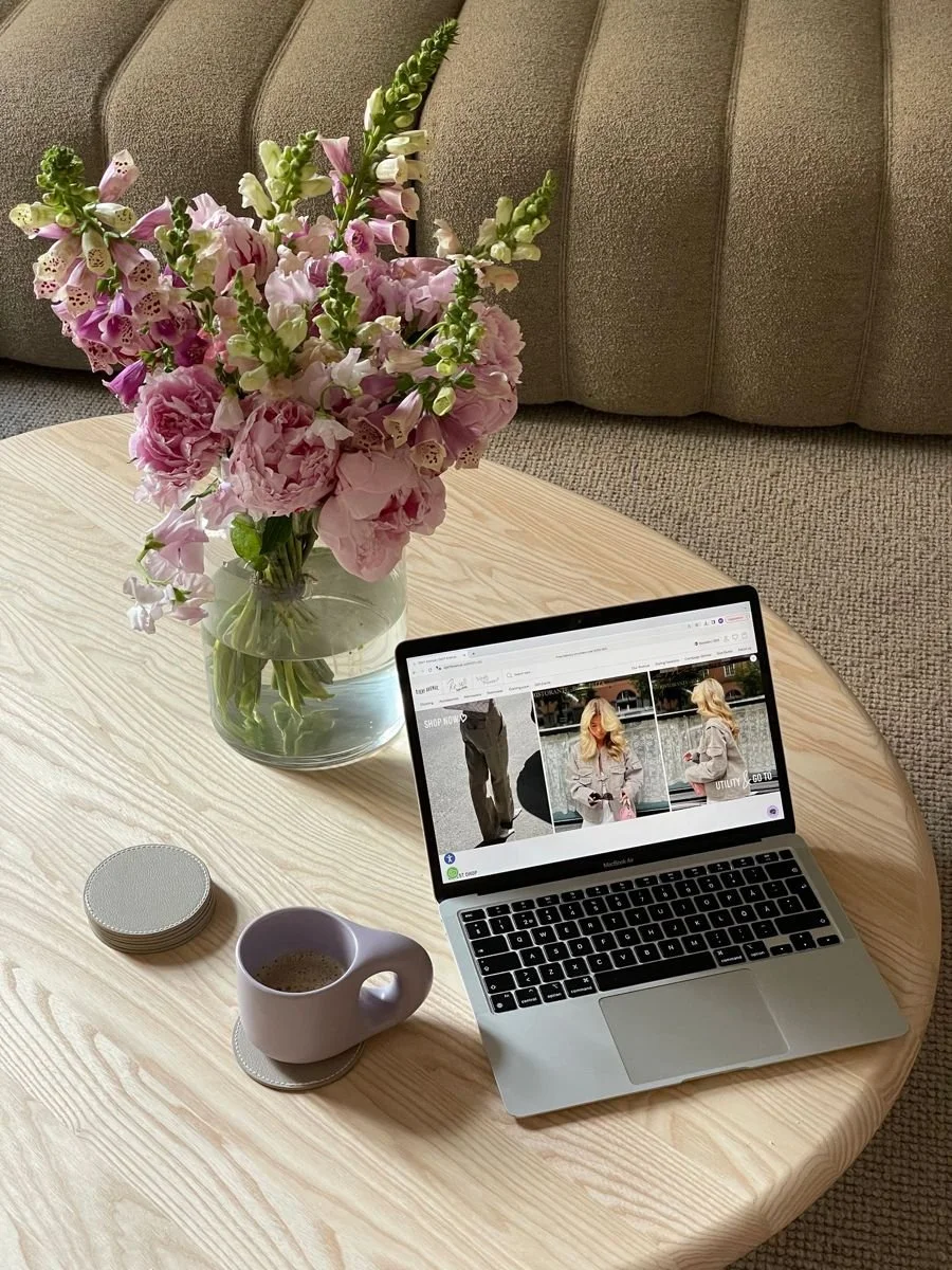 A round wooden table with a pink floral arrangement in a glass vase, a closed stack of coasters, a mug filled with coffee on a coaster, and an open MacBook displaying a webpage, in front of a beige upholstered sofa.