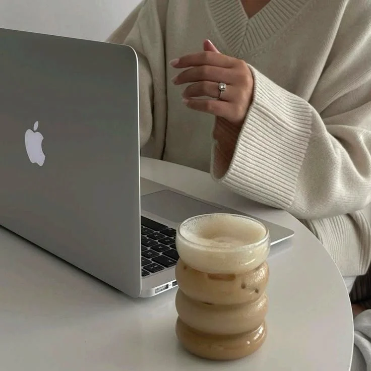 A person wearing a beige sweater sitting at a white table with a silver MacBook and a stack of glazed donuts.