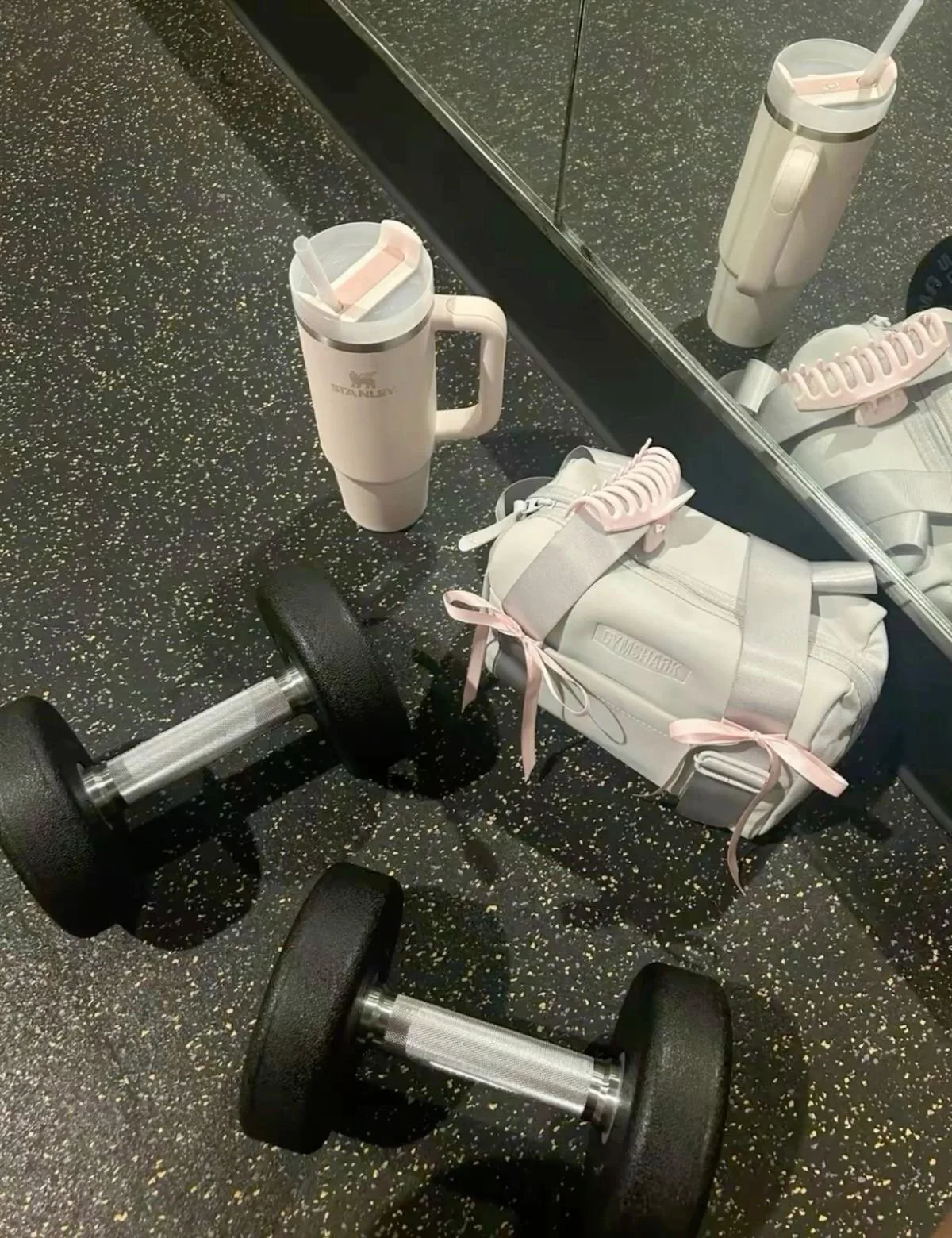 A gym floor with two dumbbells, a white gym bag with pink ribbons, a pink and white tumbler with a straw, and a pair of white sneakers with pink laces placed on top of the bag. There is a mirror reflecting the objects and the gym floor.