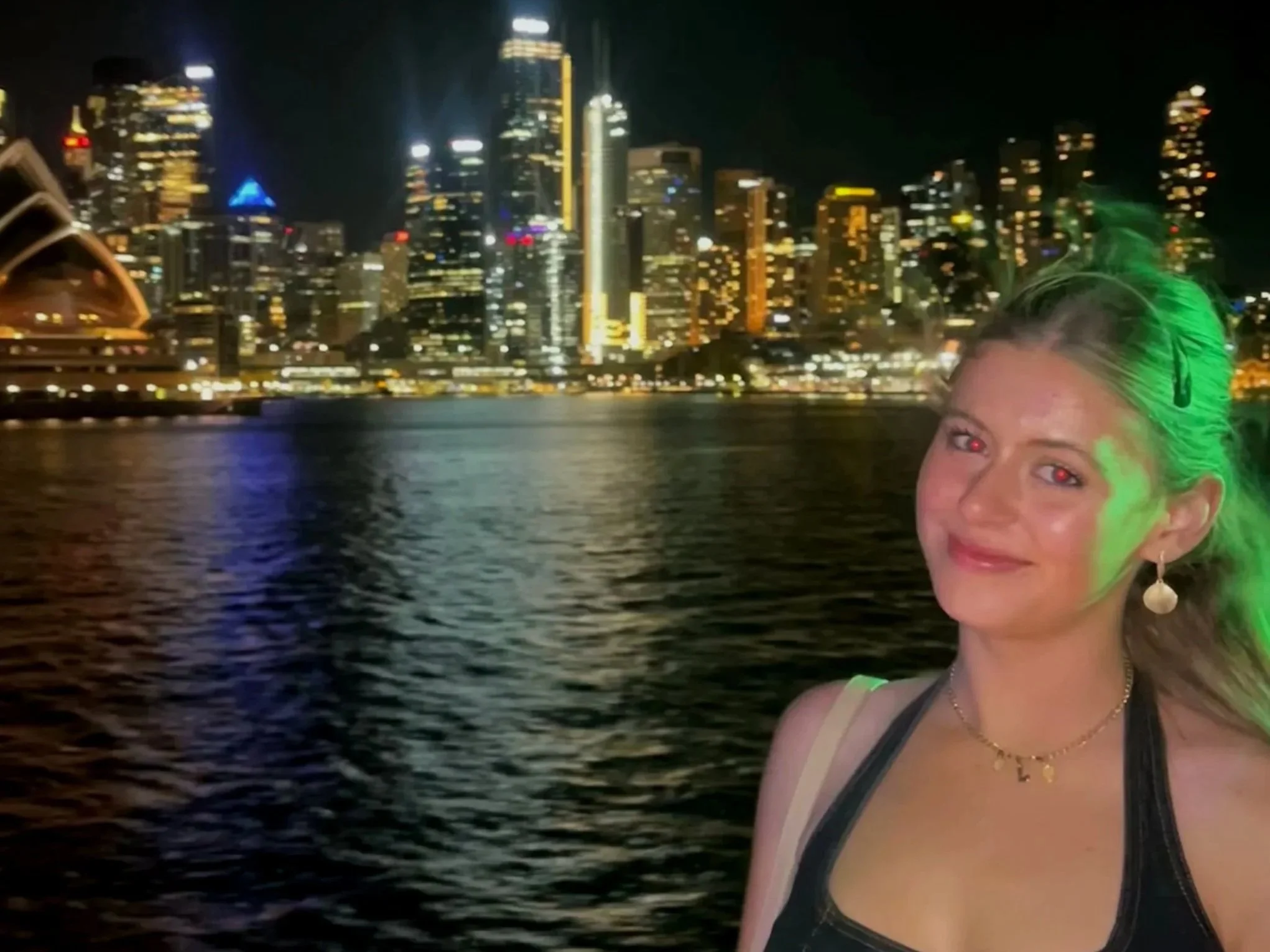 A woman standing by a body of water at night with a city skyline lit up in the background, including tall buildings and the Sydney Opera House.