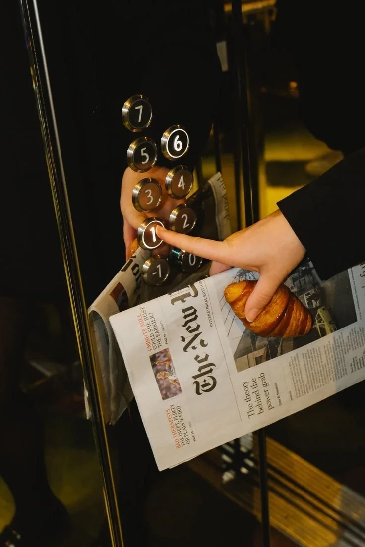 A person presses the button labeled '1' in an elevator, holding a newspaper and a croissant.