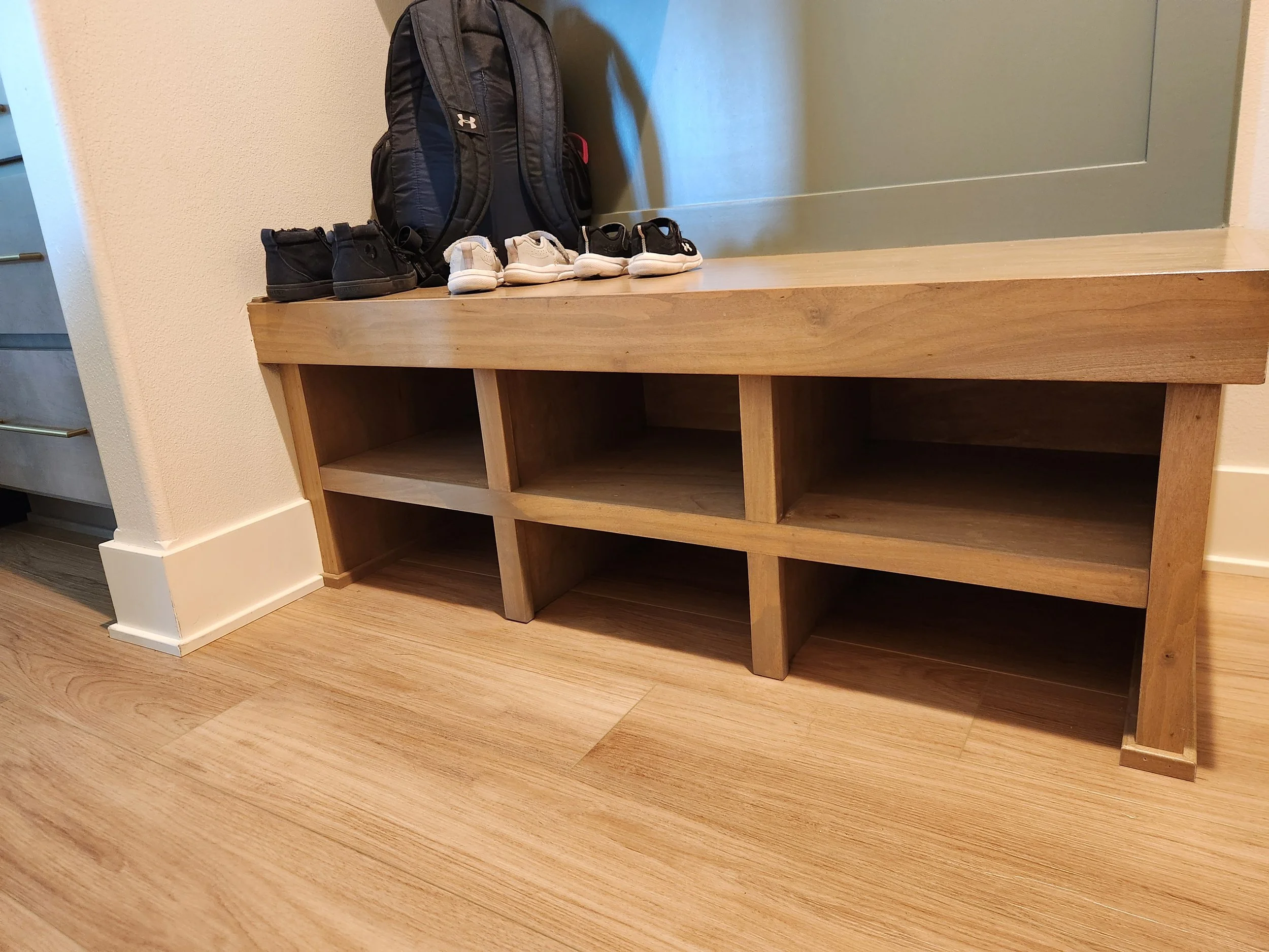 A wooden shoe rack with an open shelf design, placed against a beige wall with a small gap showing a corner. On top of the rack, there are four pairs of shoes and a black backpack leaning against the wall, with the shoes neatly arranged in pairs.