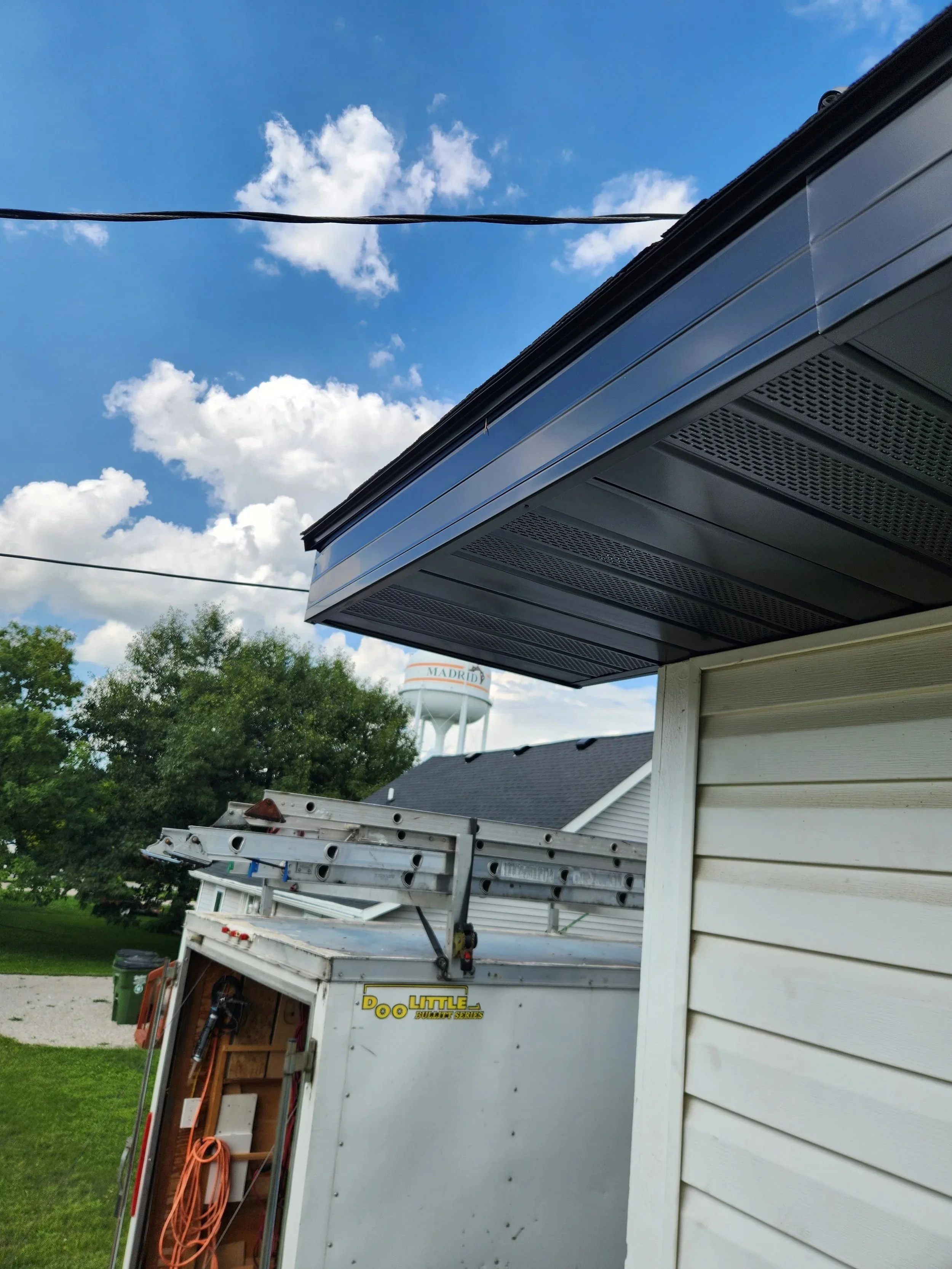 The image shows a part of a house with a corner of the roof and siding, a utility trailer with tools and extension cords, and a water tower with the word 'MADRID' in the background under a bright blue sky with clouds.