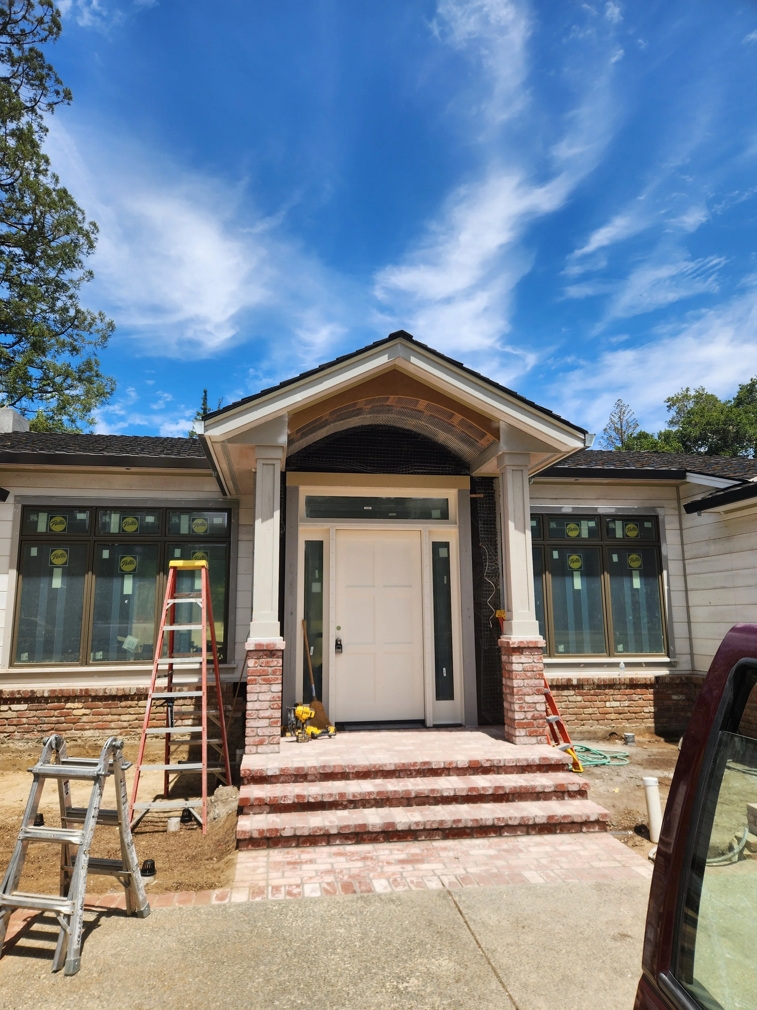 Front porch of a house under construction with brick stairs, wooden steps, and an entrance door, with construction tools and ladders present.