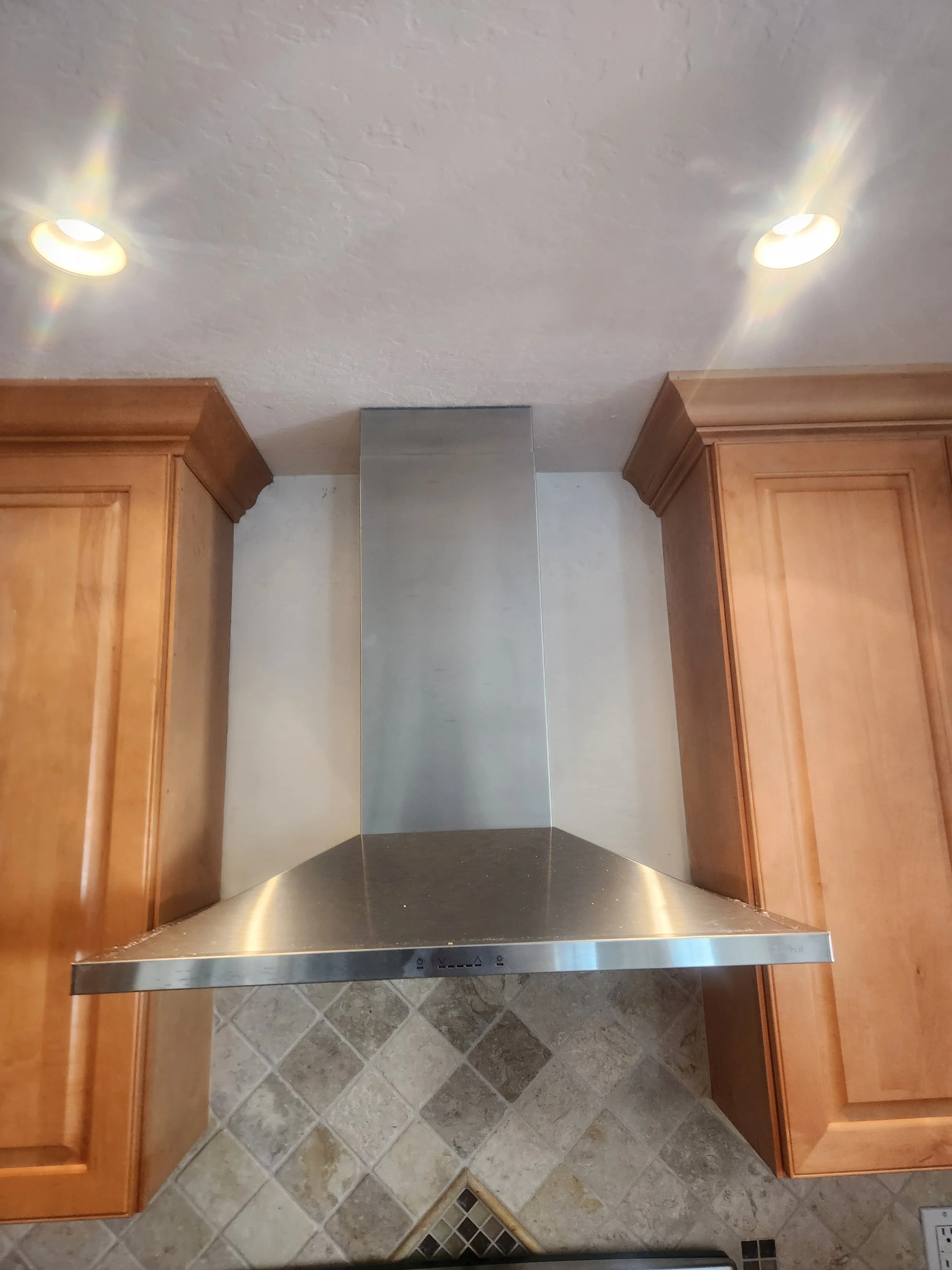 A modern kitchen range hood with a stainless steel finish installed between two wooden cabinets. The hood has control buttons on its front edge and is mounted against a white wall. The ceiling has two recessed lights, and the floor is tiled with a beige and brown pattern.