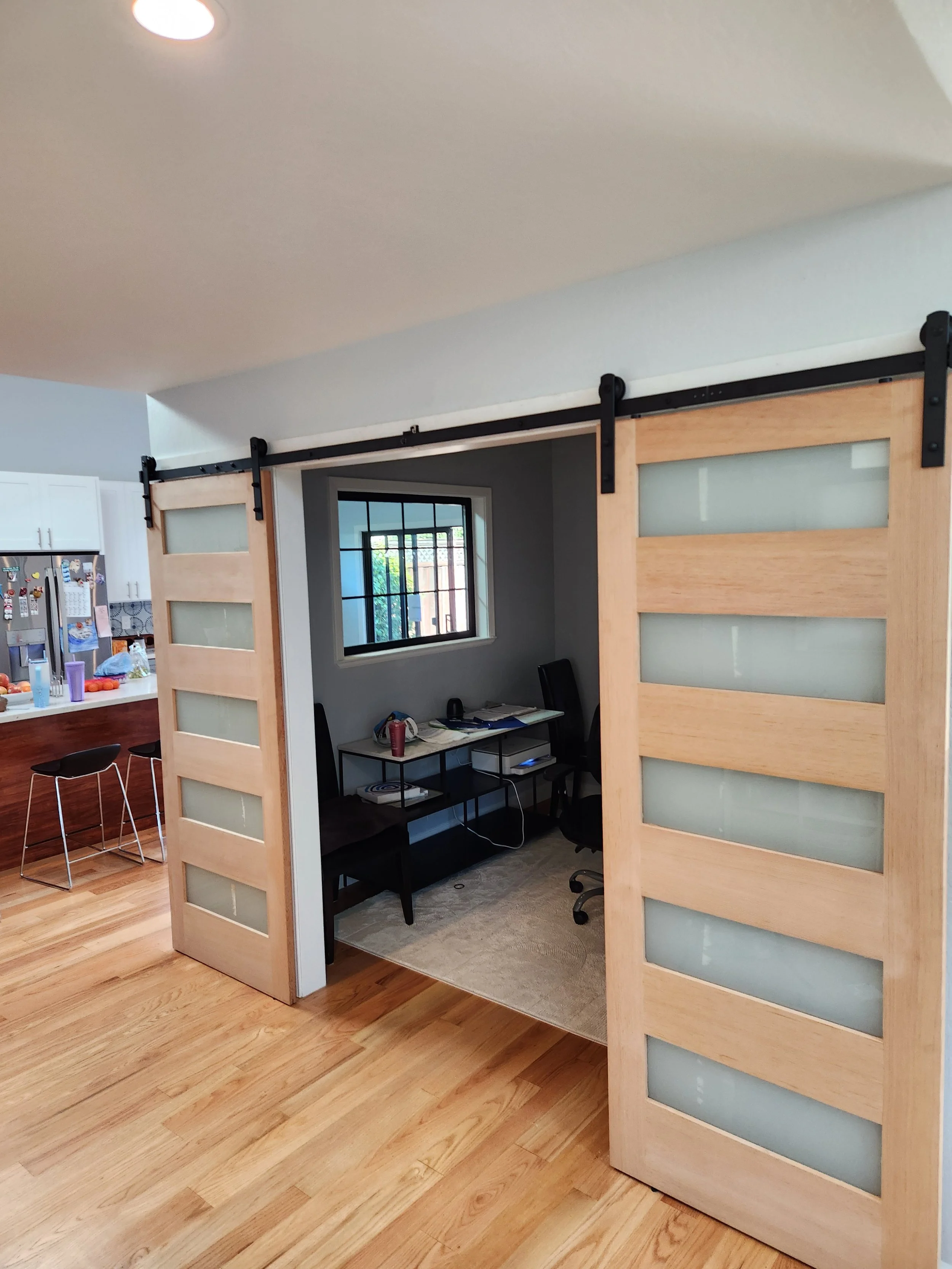 A sliding barn door made of light colored wood with frosted glass panels, mounted on black metal hardware, partially open to reveal a small office with a black desk, two black chairs, a window, and some papers. The room has light gray walls and a beige carpet.