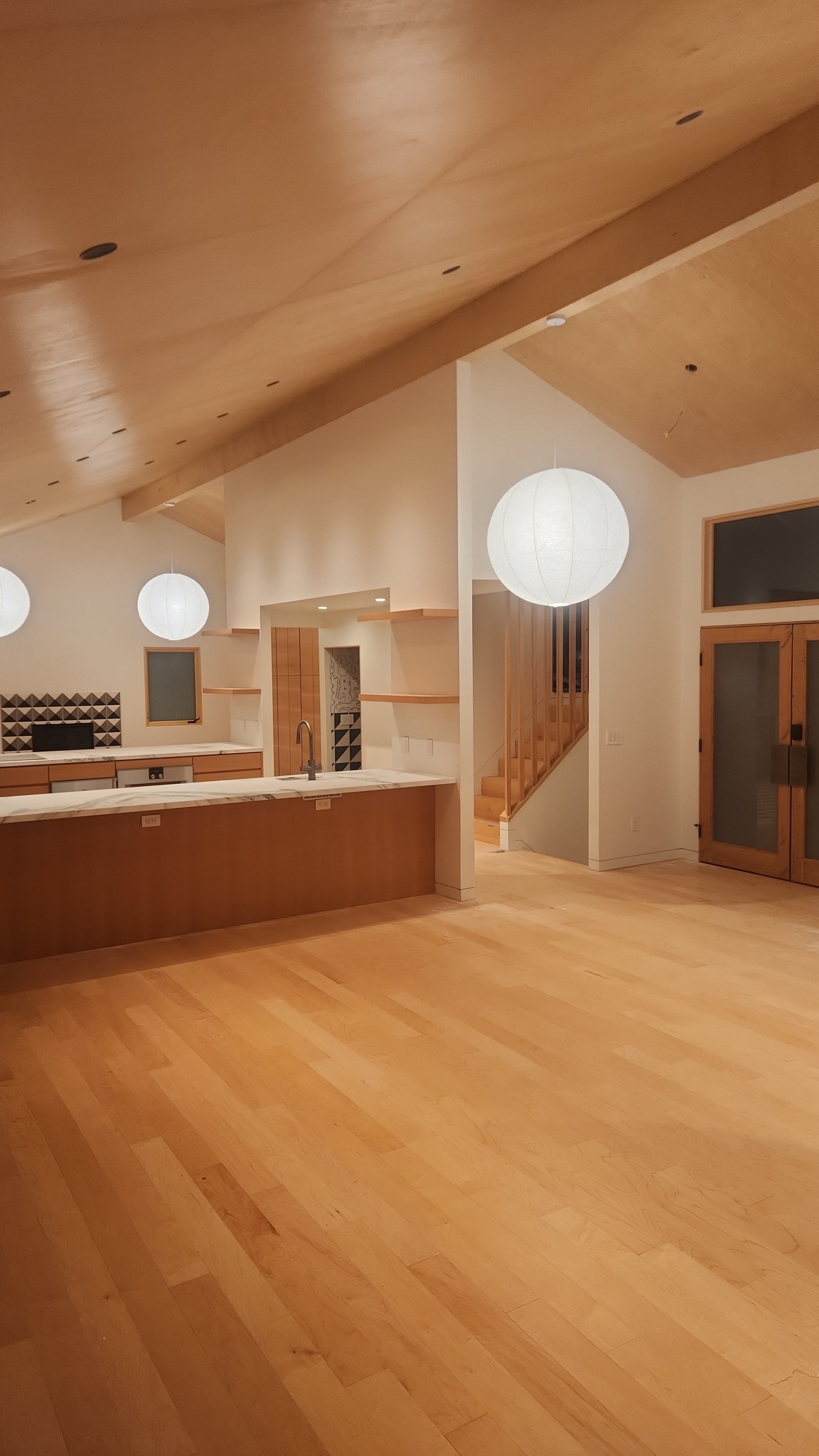 Interior view of a modern, minimalist kitchen with wooden floors, a marble-topped island, and spherical paper lantern pendant lights. There are wooden cabinets, small windows, and a staircase leading to an upper level.