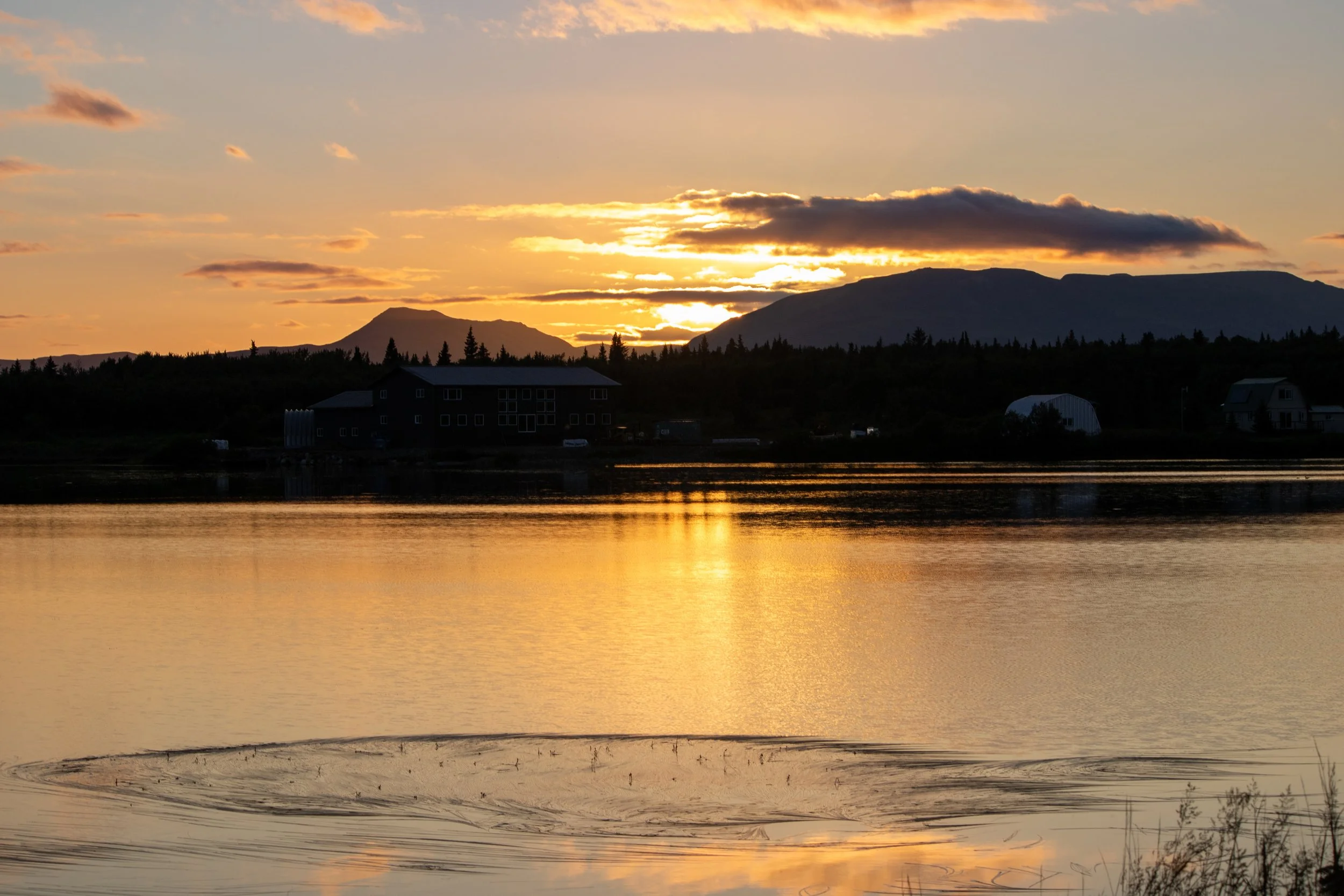 Sunset over mountains reflecting on a calm lake with houses and trees in the background.