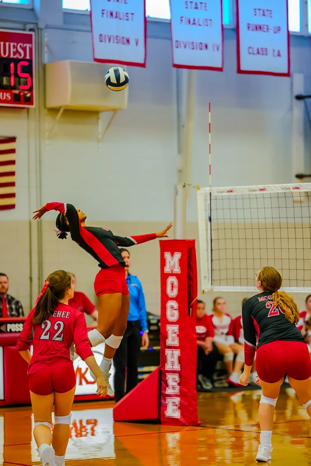 A volleyball game in progress at a gymnasium, with players in red and black uniforms. One player is jumping near the net to hit the ball while following through with her arm. Other players and spectators are visible in the background.