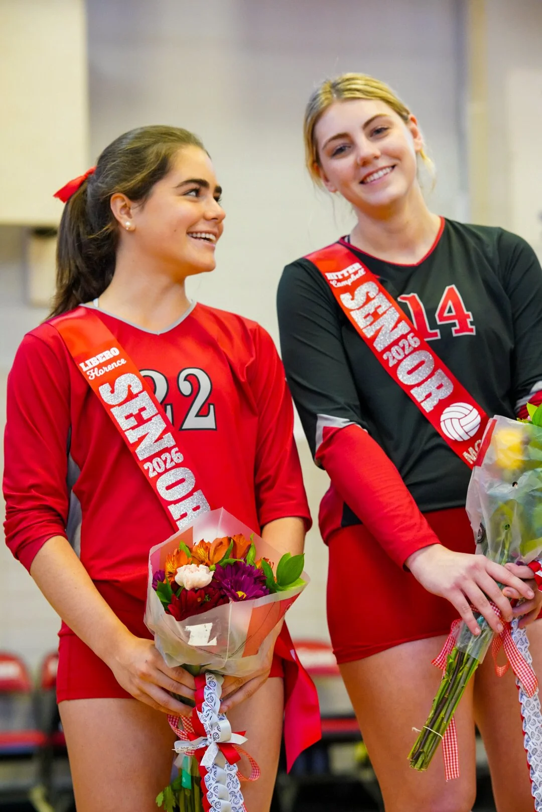 Two young female volleyball players in red and black uniforms holding flower bouquets, smiling, with sashes indicating event participation in 2026.