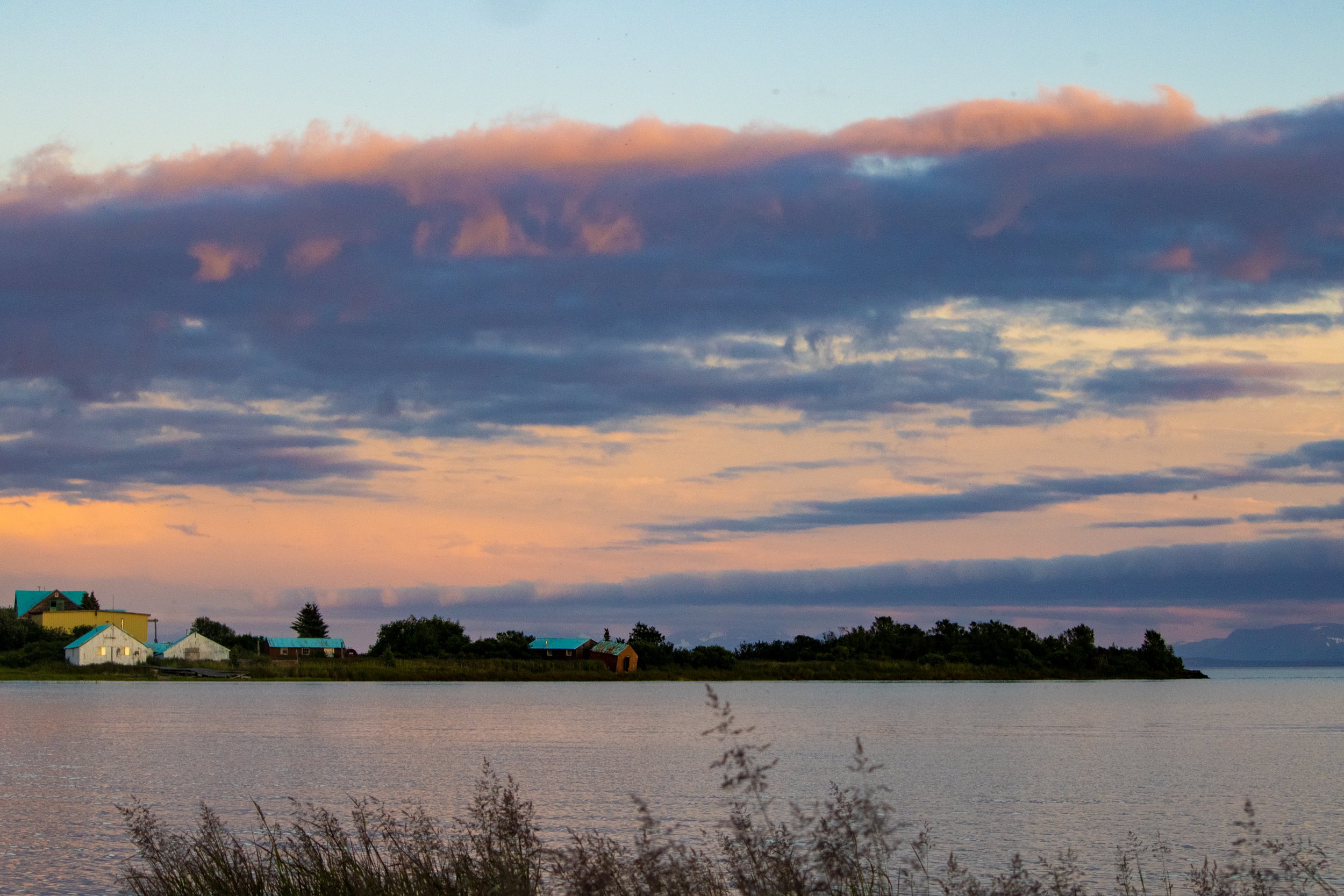 A serene lakeside scene at sunset with colorful clouds in the sky and small houses with blue roofs along the shoreline, with tall grasses in the foreground.