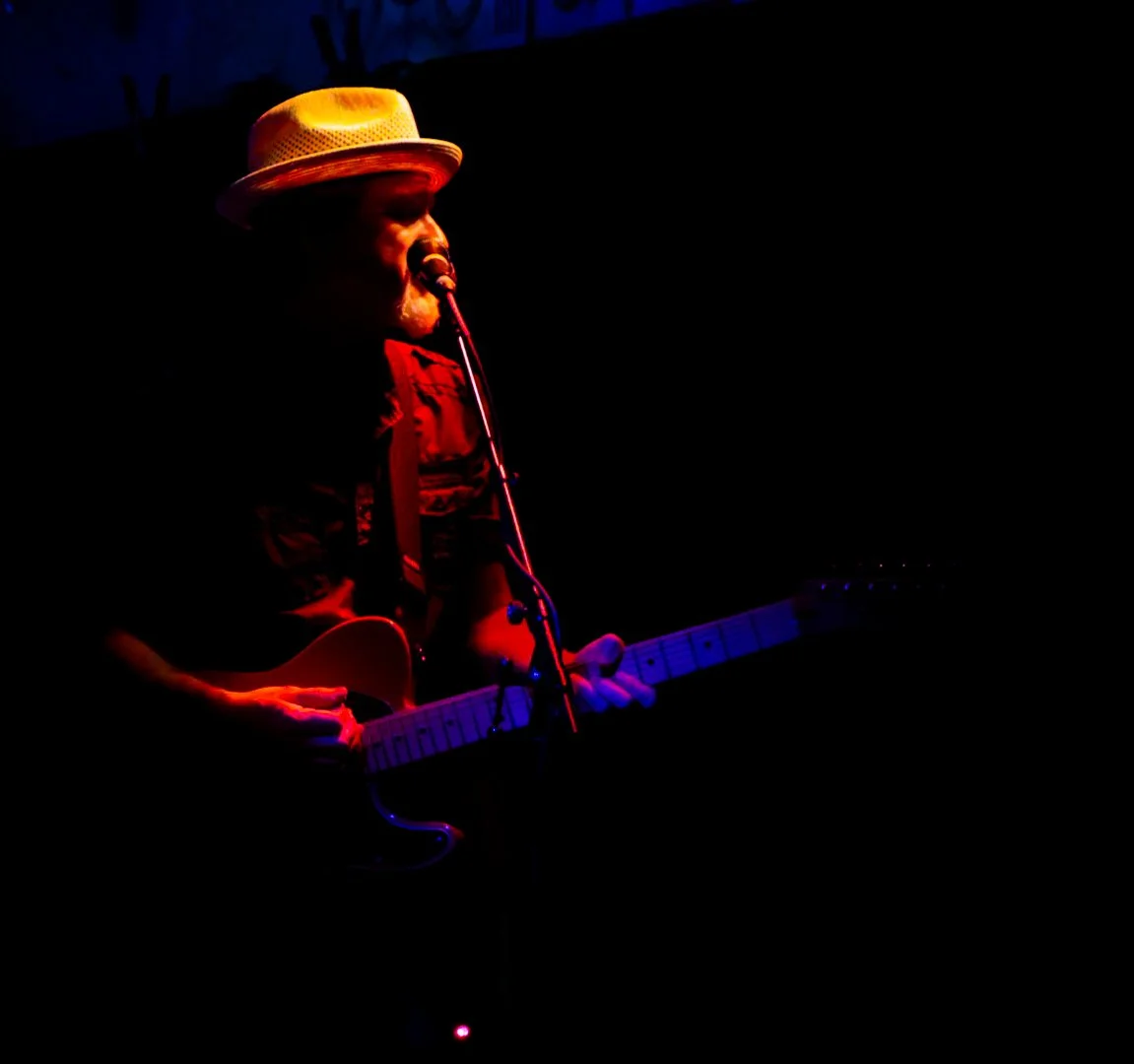 A musician wearing a straw hat and red shirt playing an electric guitar on a dark stage, illuminated by red and purple lighting.