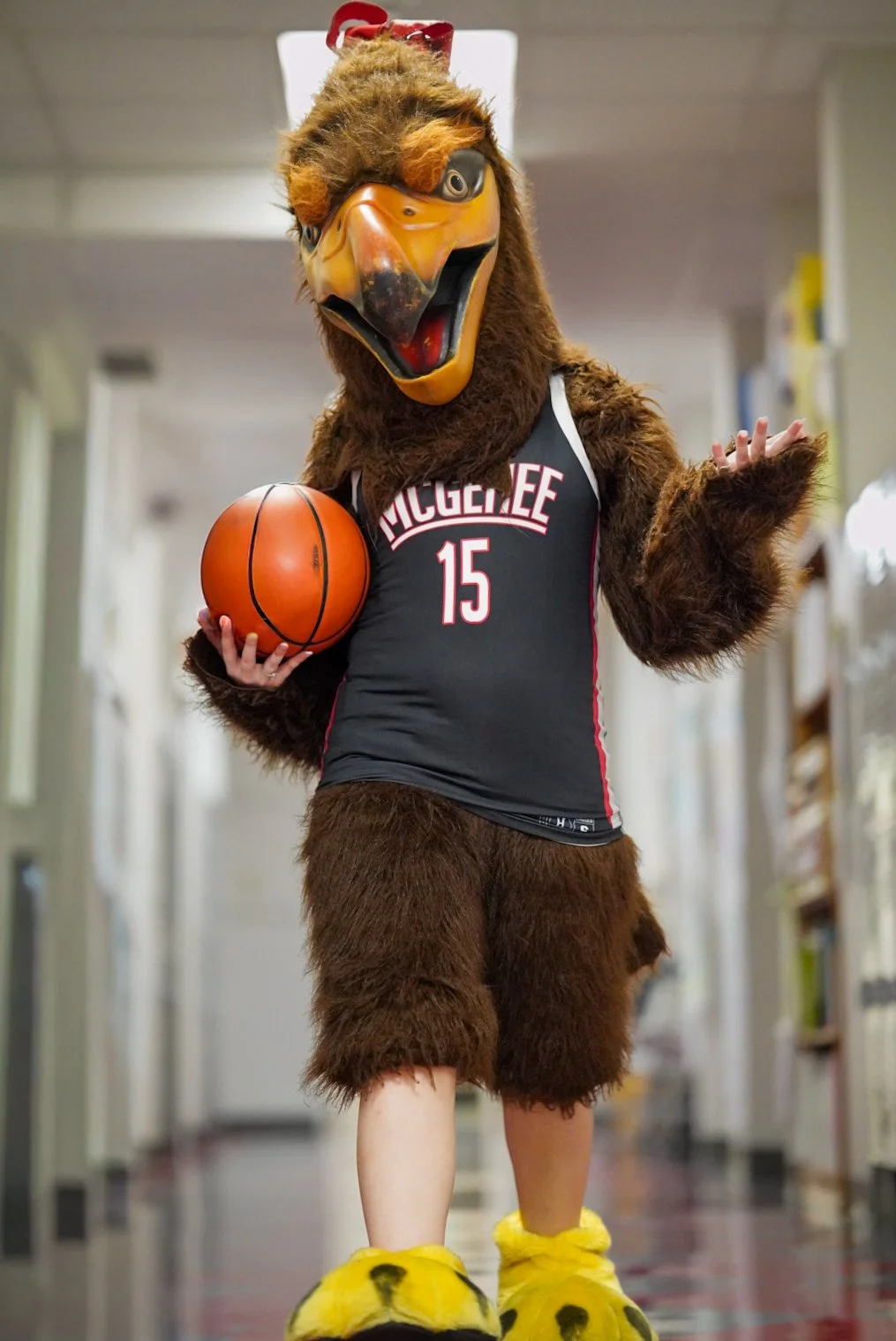 A person dressed in a large bird mascot costume with a beak and feathers, wearing a black sports jersey with the number 15 and the word Muggee, holding a basketball, standing in a hallway.