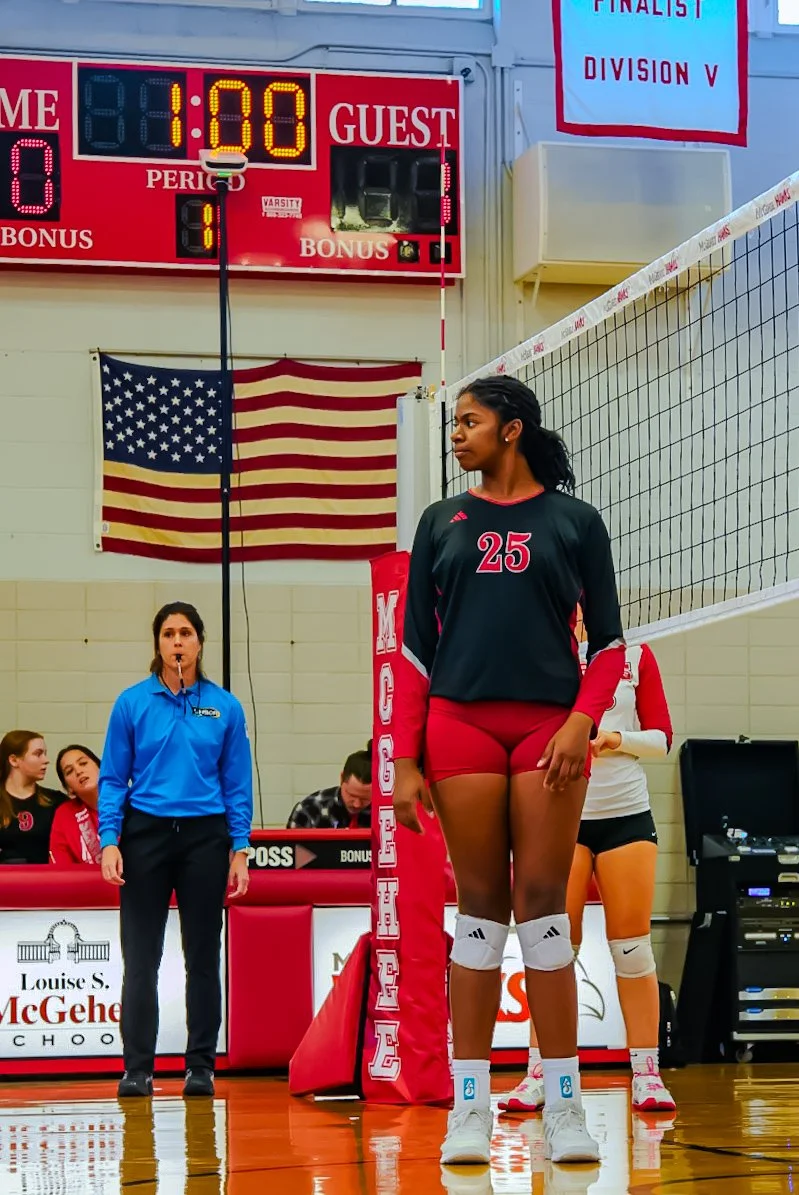 A female volleyball player standing on the court in a black and red uniform with the number 25, with a volleyball net and referee in the background, during a match in a gymnasium with an American flag and a scoreboard showing 1:00 minutes left.
