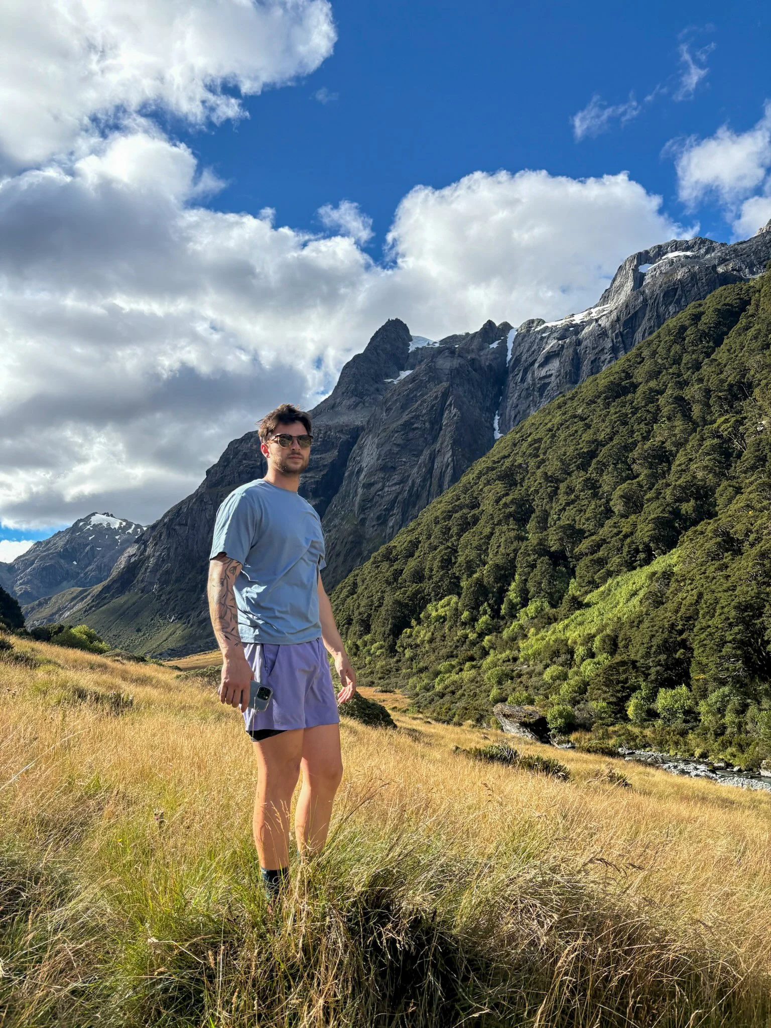 A man standing in a grassy valley with tall rocky mountains and a partly cloudy sky in the background.