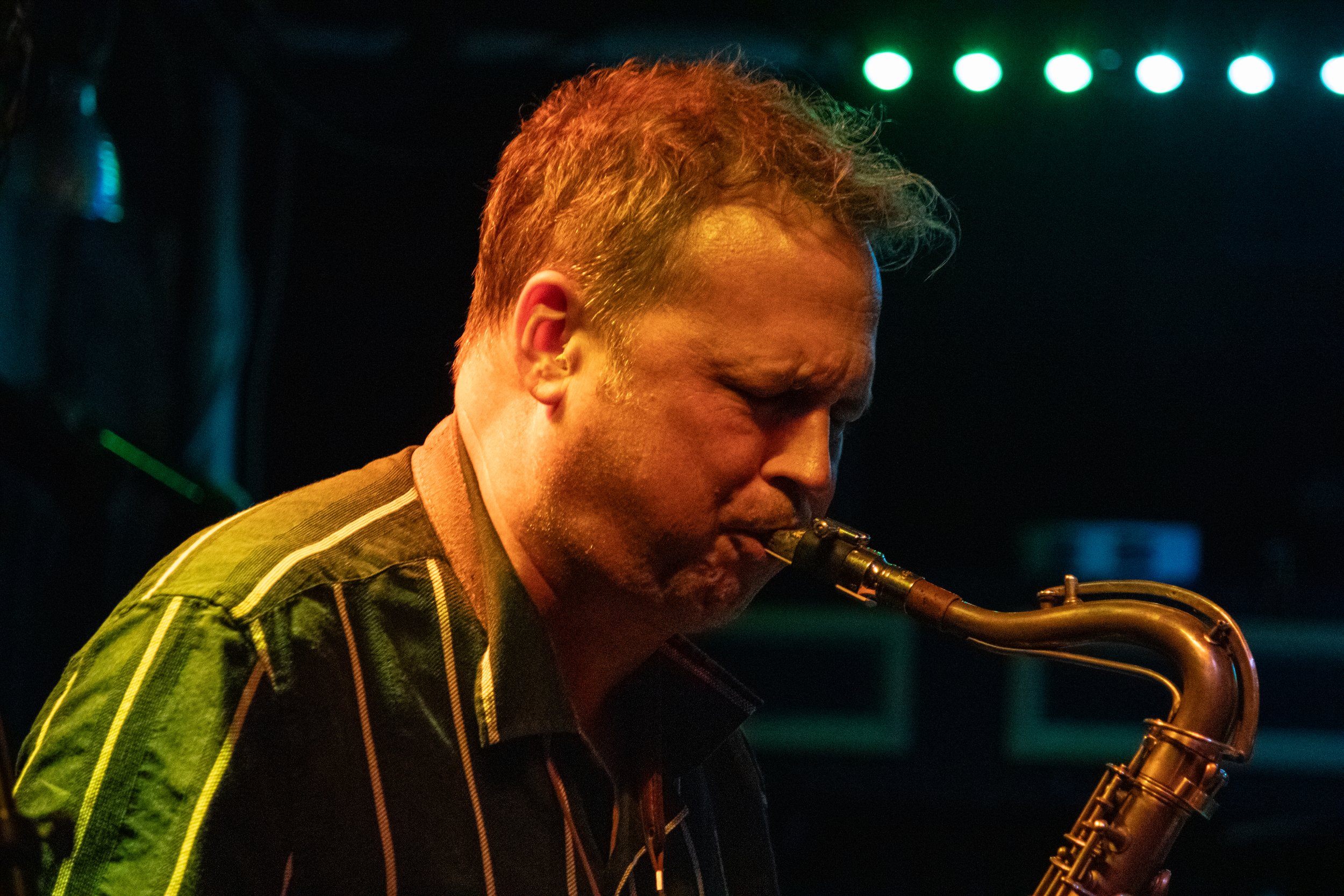 Man playing saxophone on stage in dim lighting with green and blue stage lights in the background.
