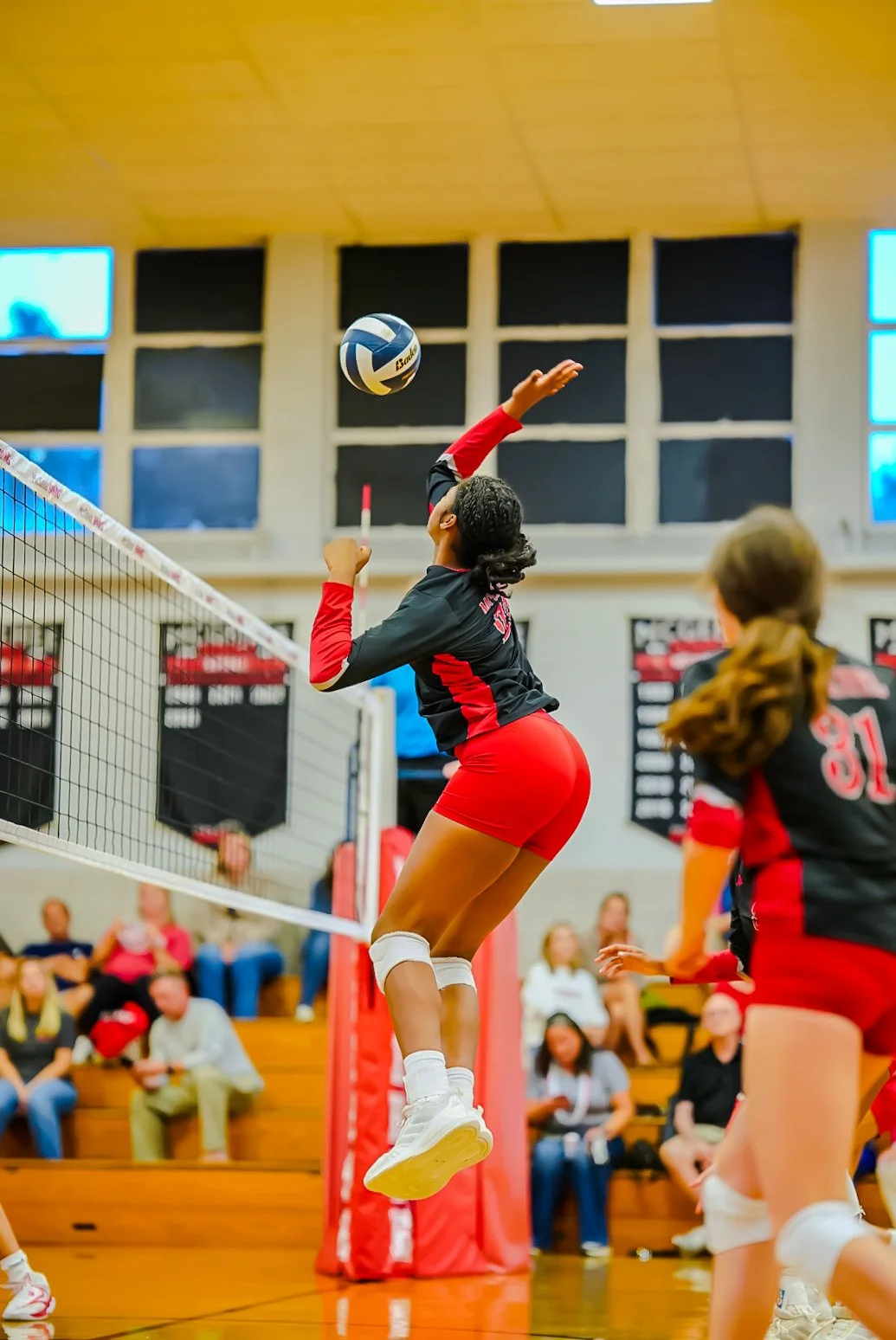 A volleyball player in a black and red uniform jumps to hit the ball at the net during a match in a gymnasium, with spectators watching in the background.