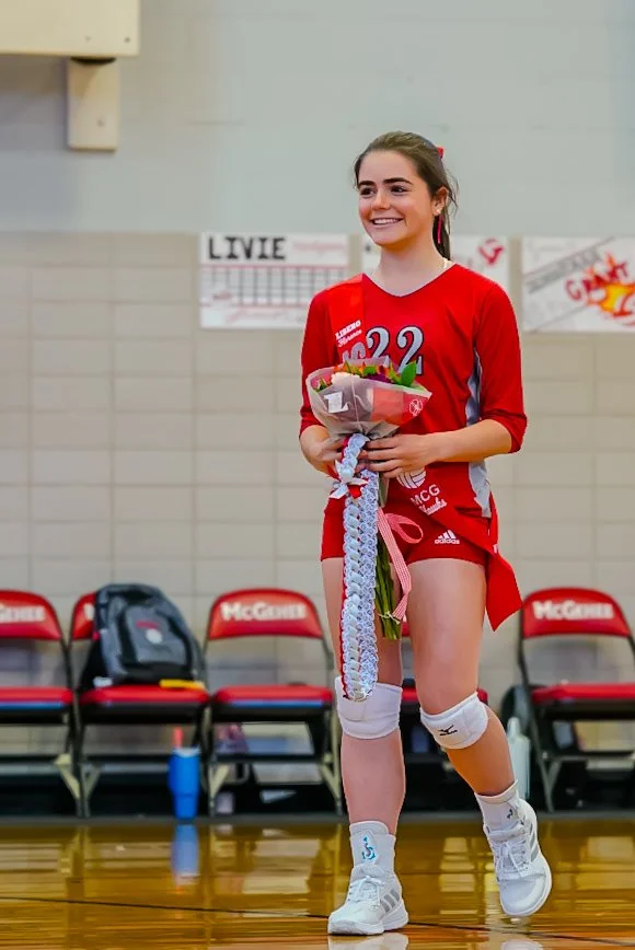 Young female volleyball player in red uniform holding a bouquet of flowers, standing on a gym court, smiling.