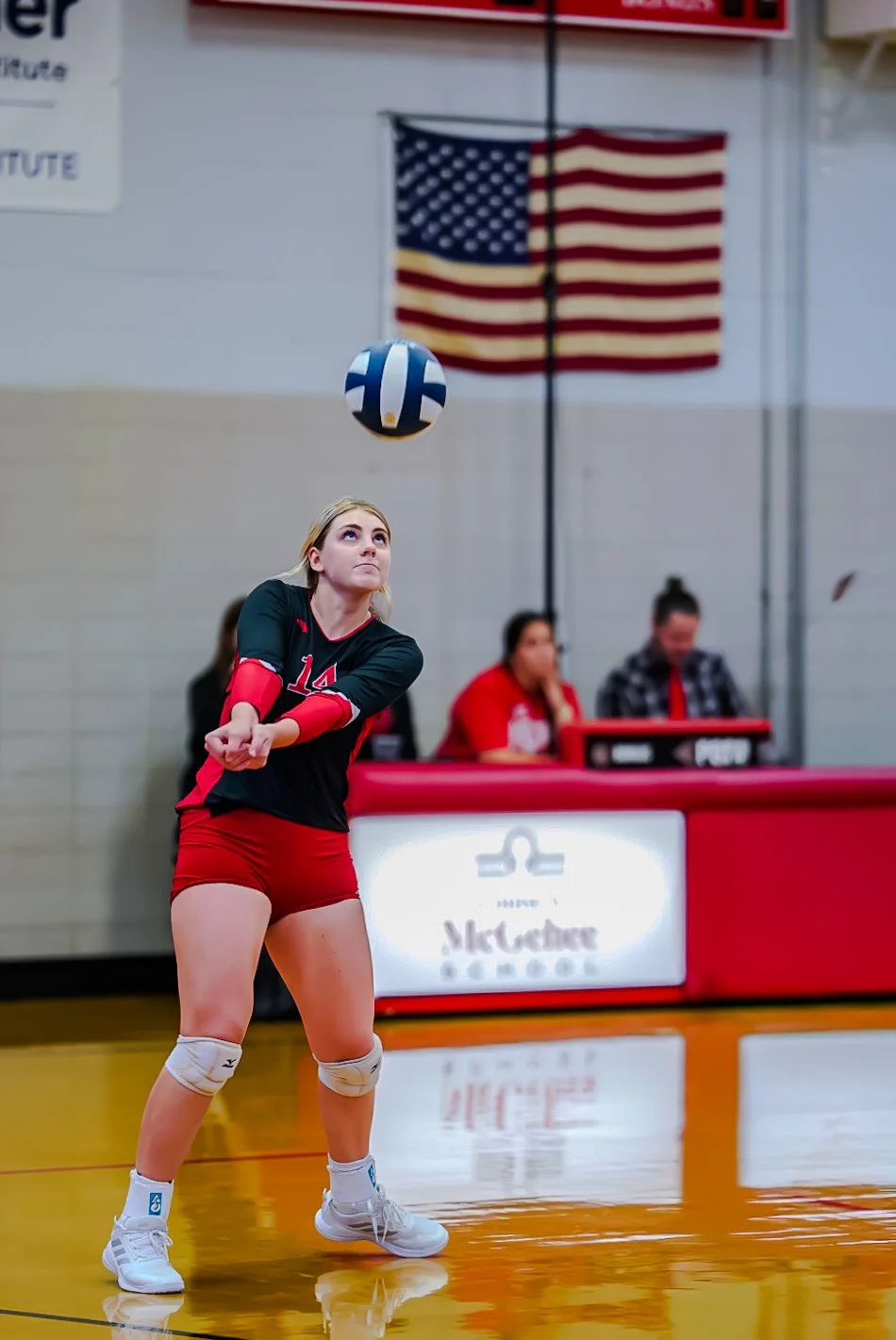 A female volleyball player in a black and red uniform hitting a volleyball inside a gymnasium, with an American flag in the background.