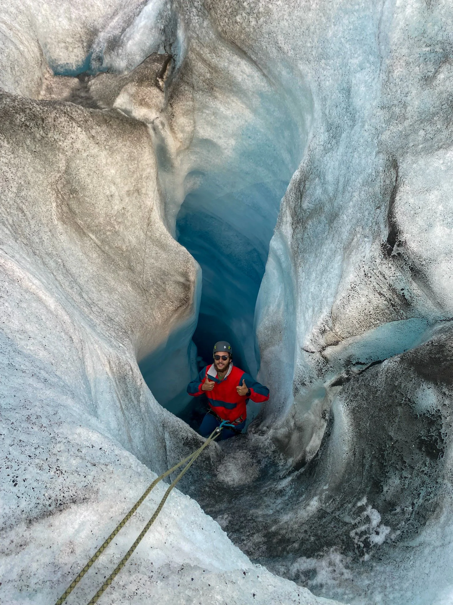 A person in climbing gear, including a helmet and red jacket, stands inside a narrow glacier crevasse with ice walls and blue ice formations, giving a thumbs-up.