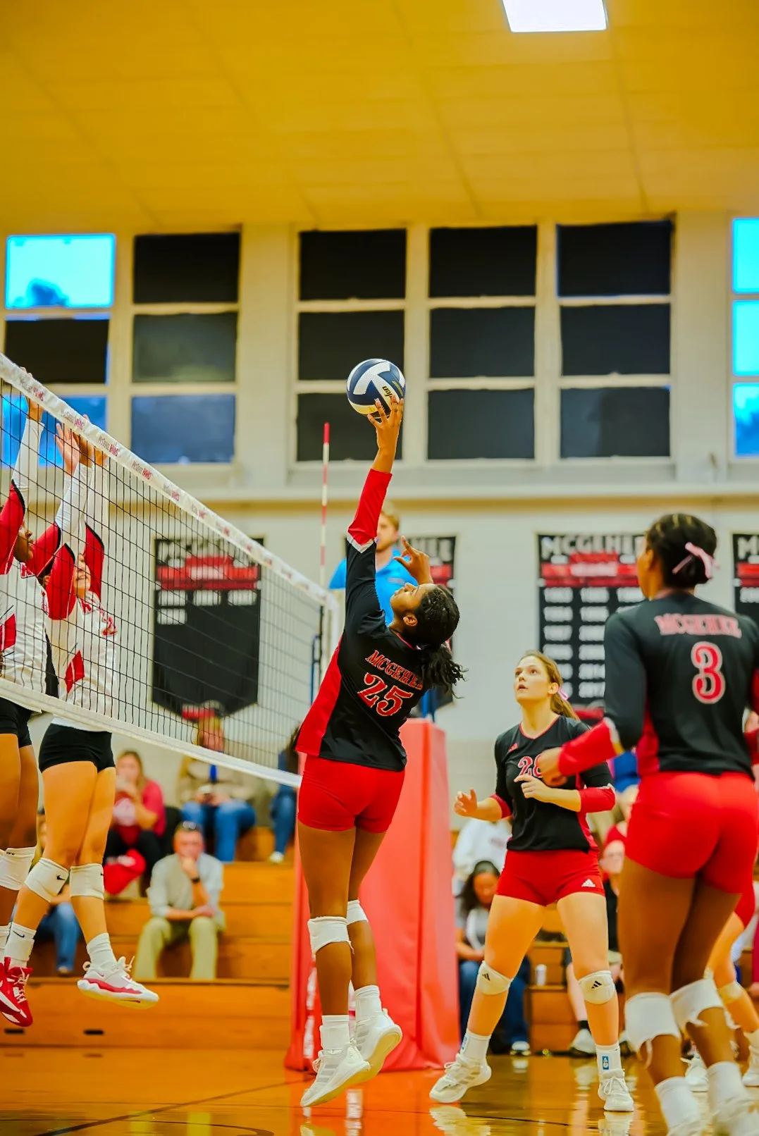 Girls volleyball game in progress, with one player jumping to hit the ball over the net while her teammates watch nearby.