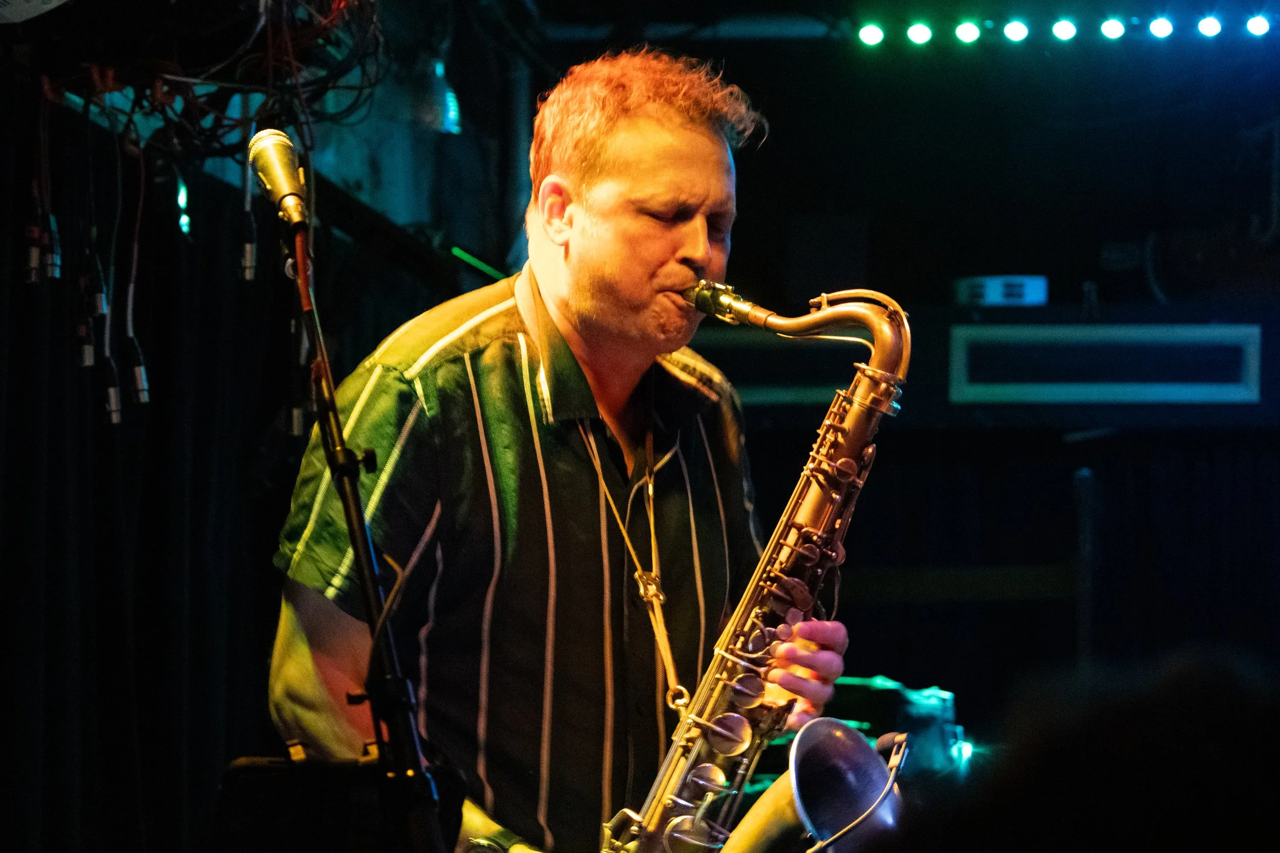 A man with short reddish hair playing a saxophone on stage, with colorful stage lighting and a dark background.