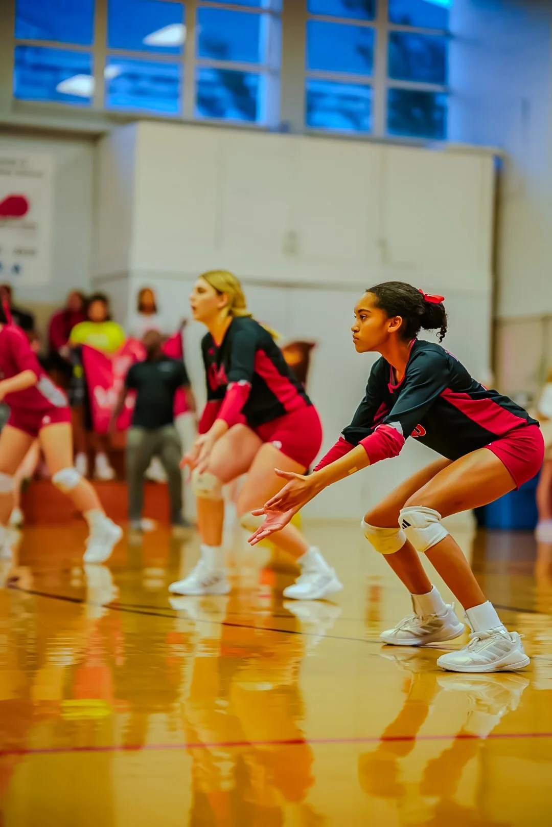 Women volleyball players in black and red uniforms performing a passing drill on a gymnasium court.