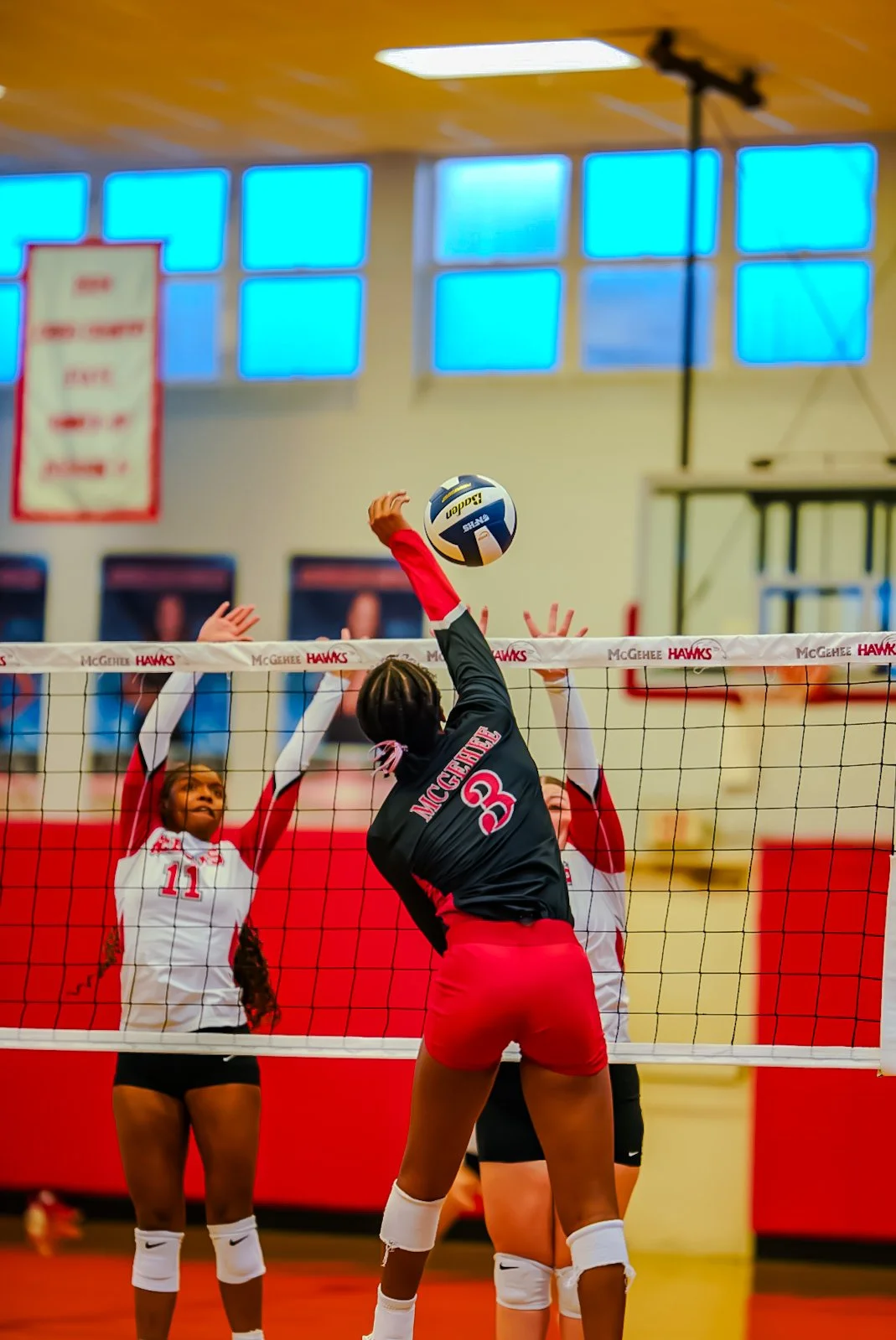 Women playing volleyball in a gymnasium, with one player jumping to hit the ball over the net, while another player on the opposite side prepares to block.