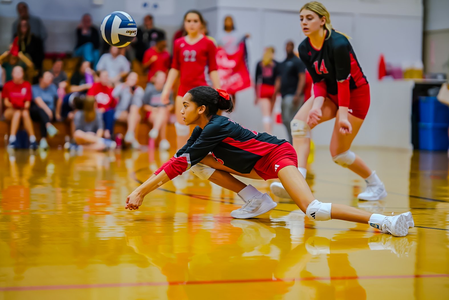A volleyball player in black and red uniform diving on the floor to save the ball during a game in a gymnasium with spectators in the background.