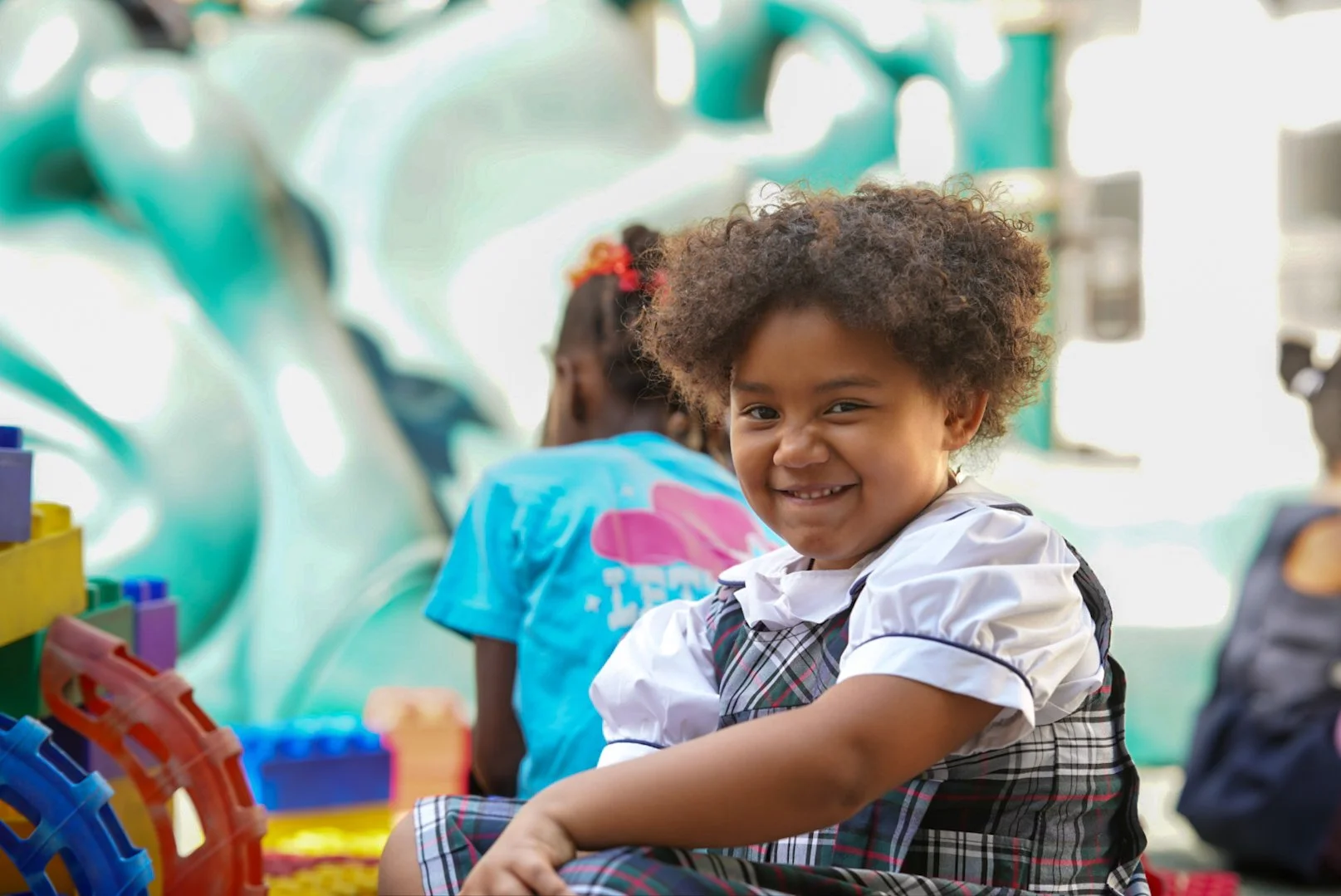 Smiling young girl in a school outfit sitting with children nearby, playing with colorful building blocks.