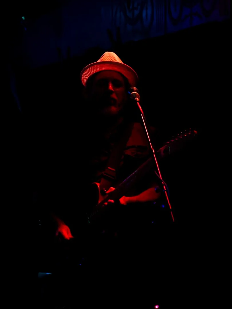 A person playing guitar on stage, wearing a hat, under red and blue lighting, with a microphone nearby.