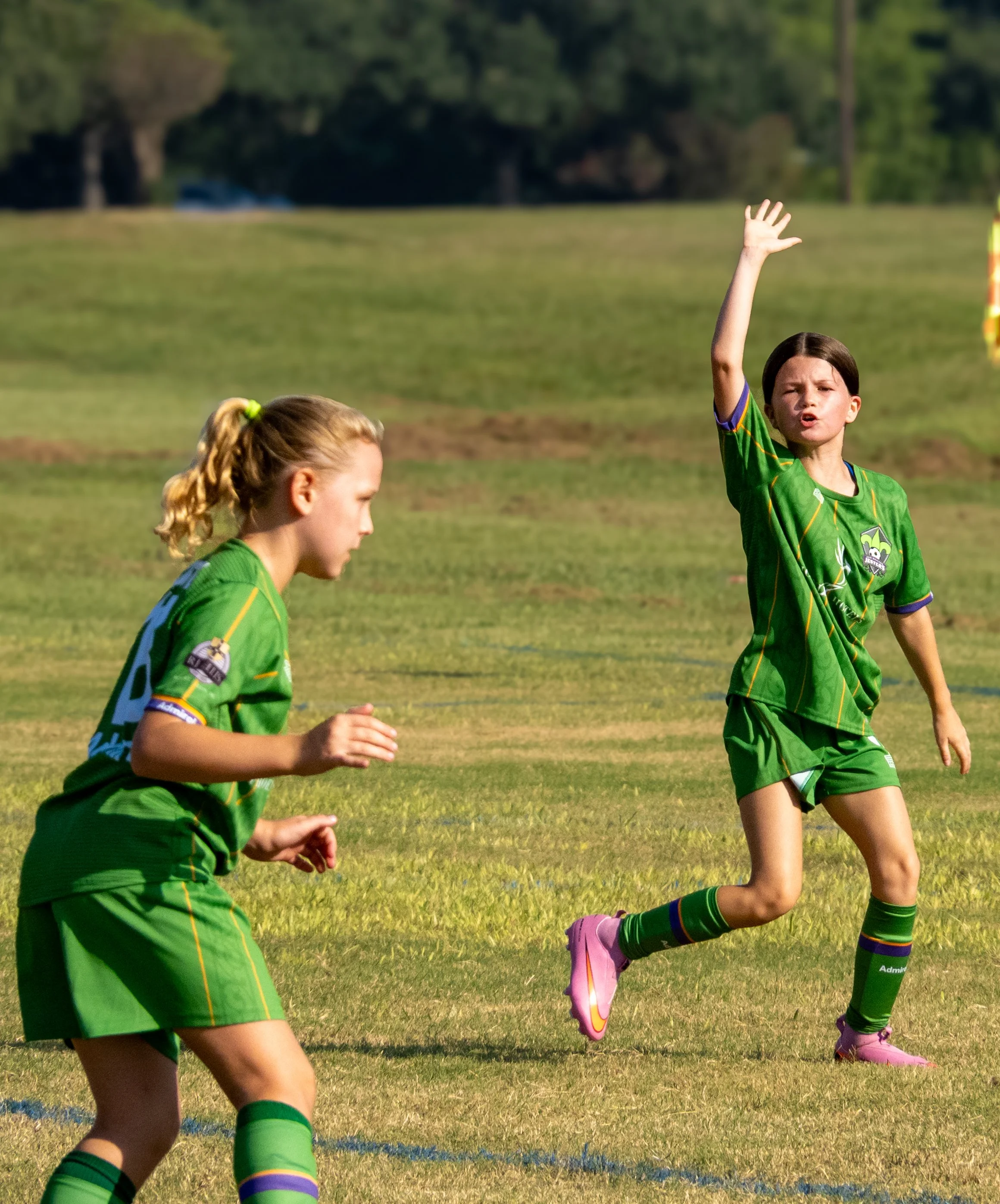 Two young girls playing soccer on a grassy field, wearing green jerseys and colorful socks. One girl is raising her hand, and the other is looking forward.