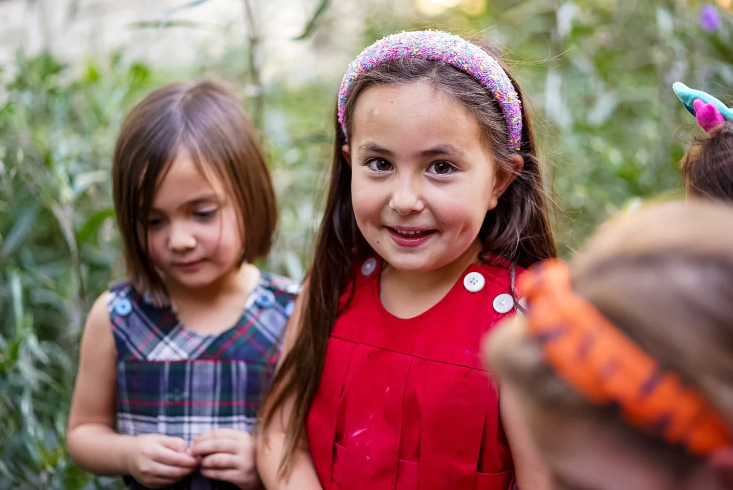 Group of young girls outdoors, one smiling at the camera with a colorful headband and red dress, others with different outfits, surrounded by greenery.