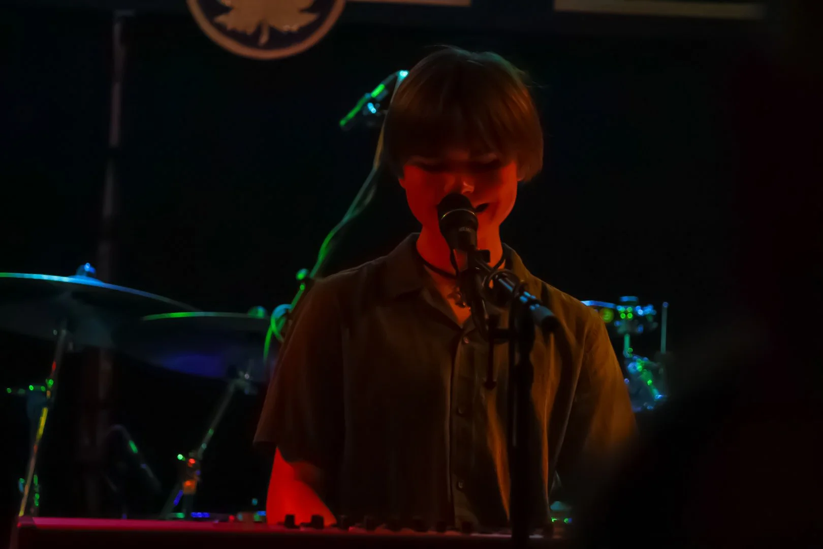 A person with short dark hair singing into a microphone on stage, illuminated by red and green stage lights, with musical equipment in the background.