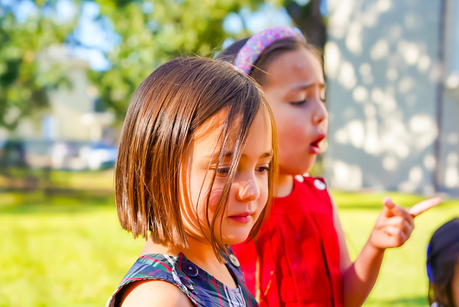 Two young girls, one with straight hair and the other with a headband, are outdoors on a sunny day, engaging with a device or object.