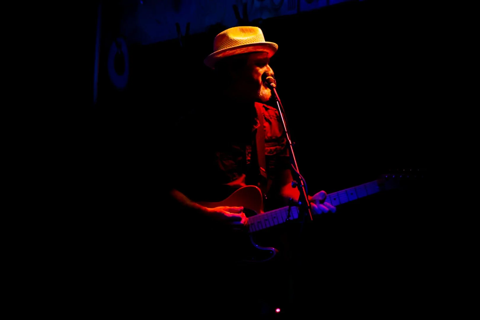 Musician wearing a straw hat and dark clothing playing an electric guitar in a dimly lit setting with minimal stage lighting.